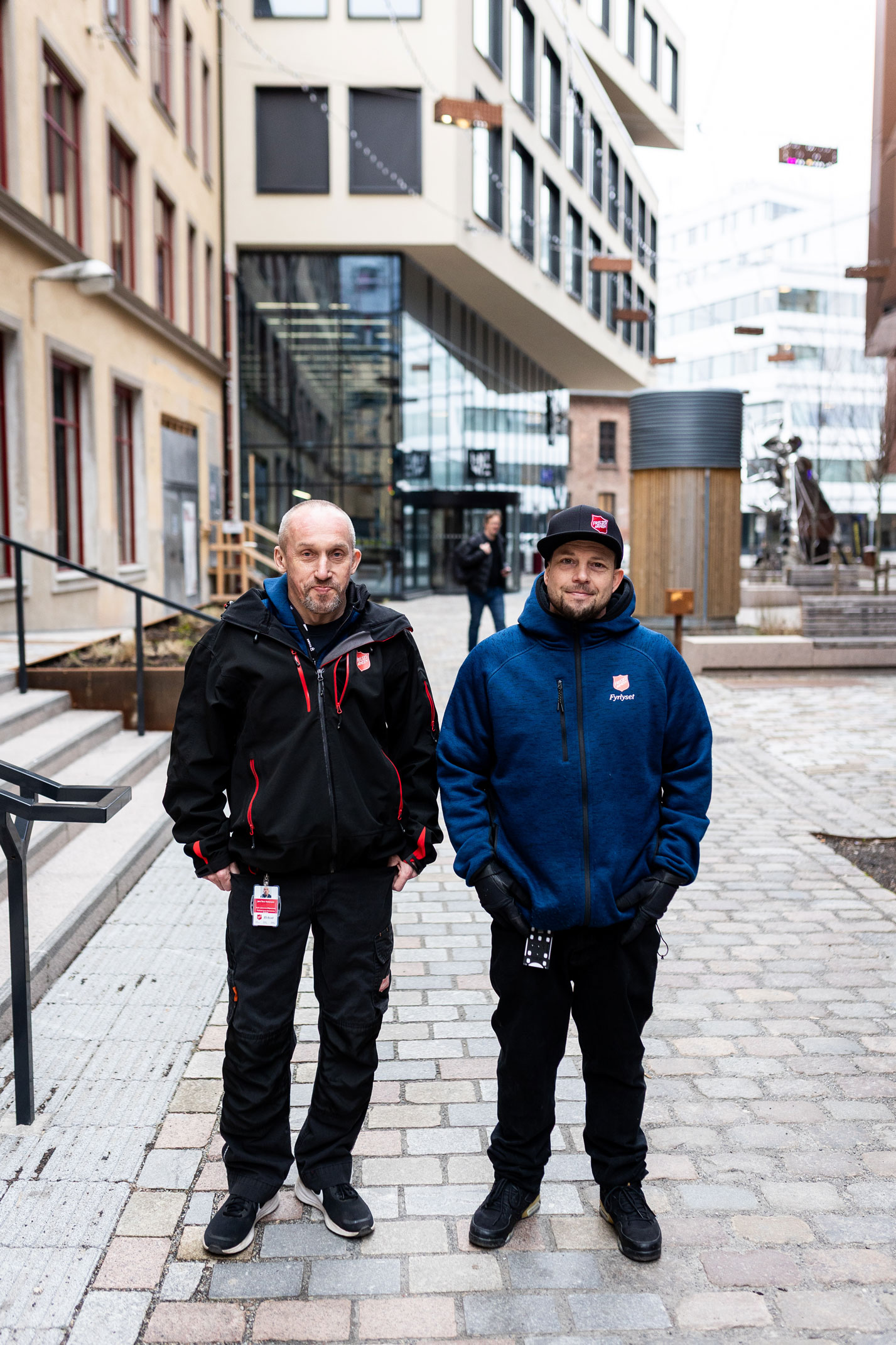 Foto: To menn med logoer fra Frelsesarmeen på klærne står i en gate med brostein. Bak dem står et nytt bygg i beige.