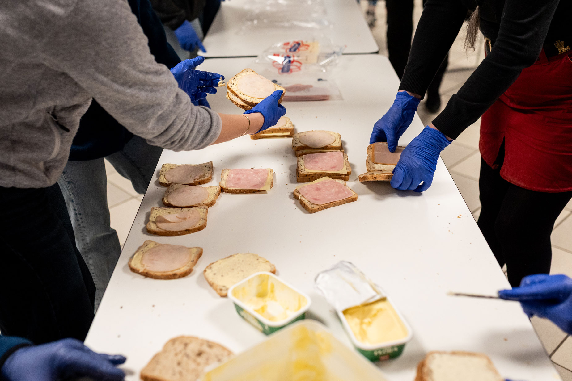 Mennsker som står rundt et bord og smører brødskiver med gulost og skinke.