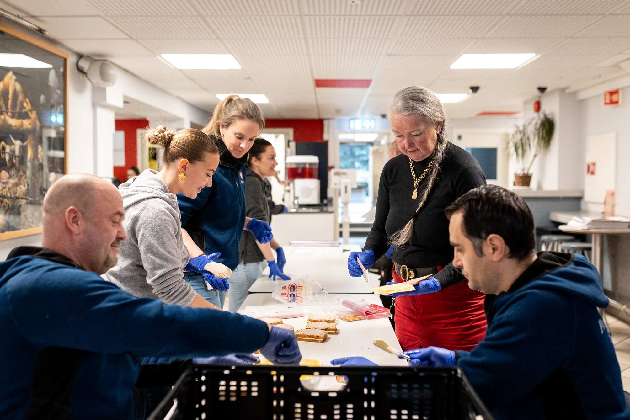 Mennsker som står rundt et bord og smører brødskiver med gulost og skinke.