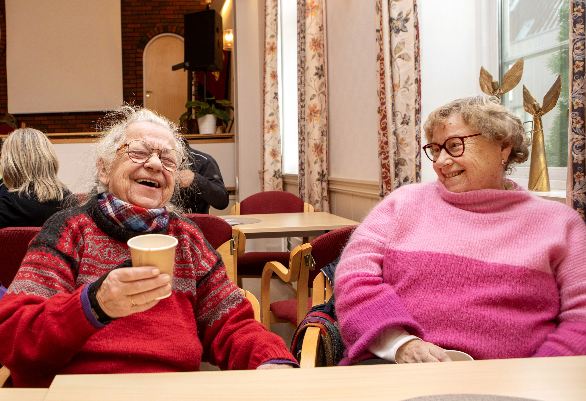 To godt voksne mennesker sitter ved et bord, drikker kaffe og smiler stort.