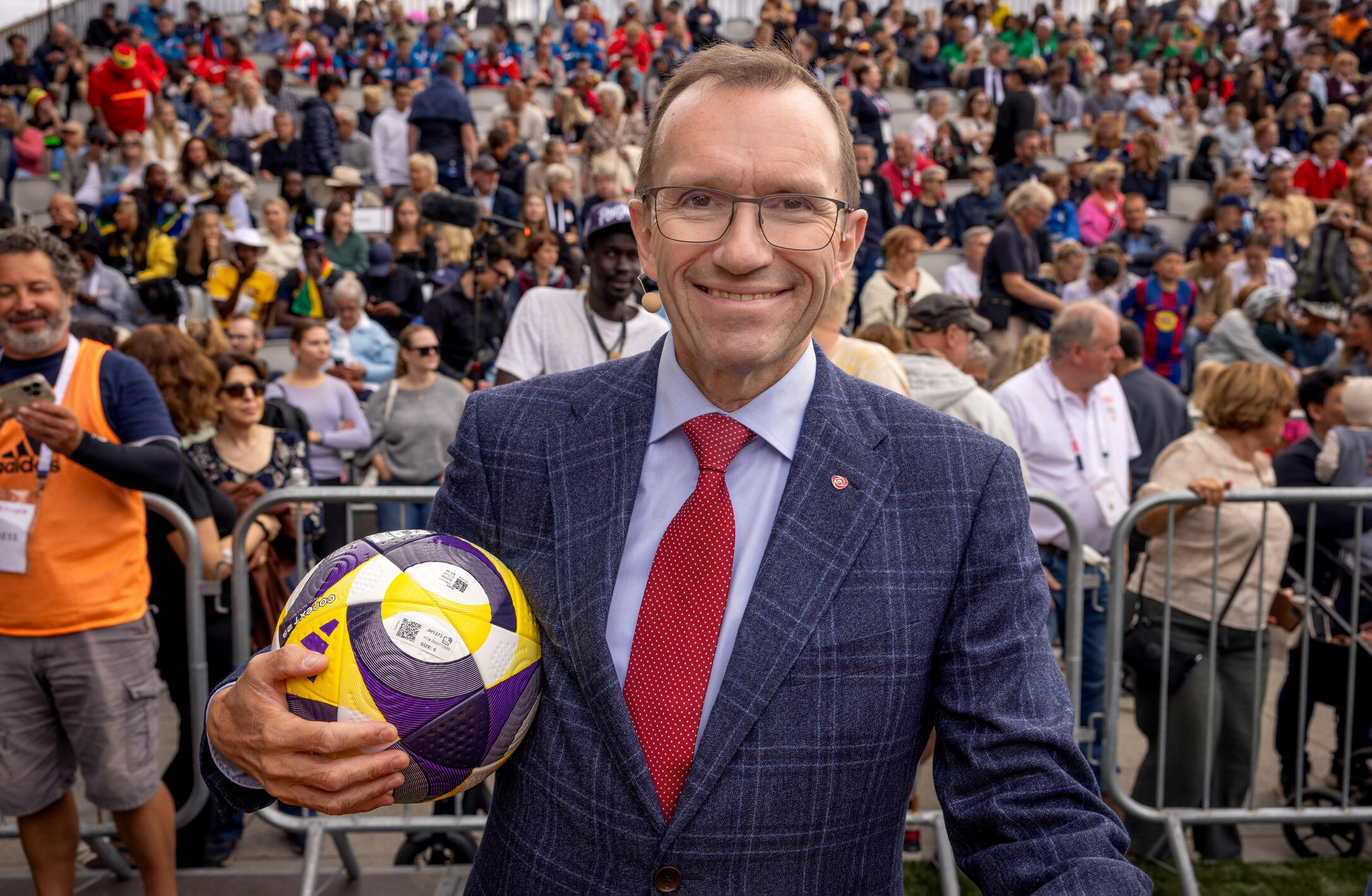 Foto: Mann i blå dress med rødt slips holder en fotball. Han har mørkt hår, litt tynt på toppen, og briller. Bak ham er en tribune full av folk.