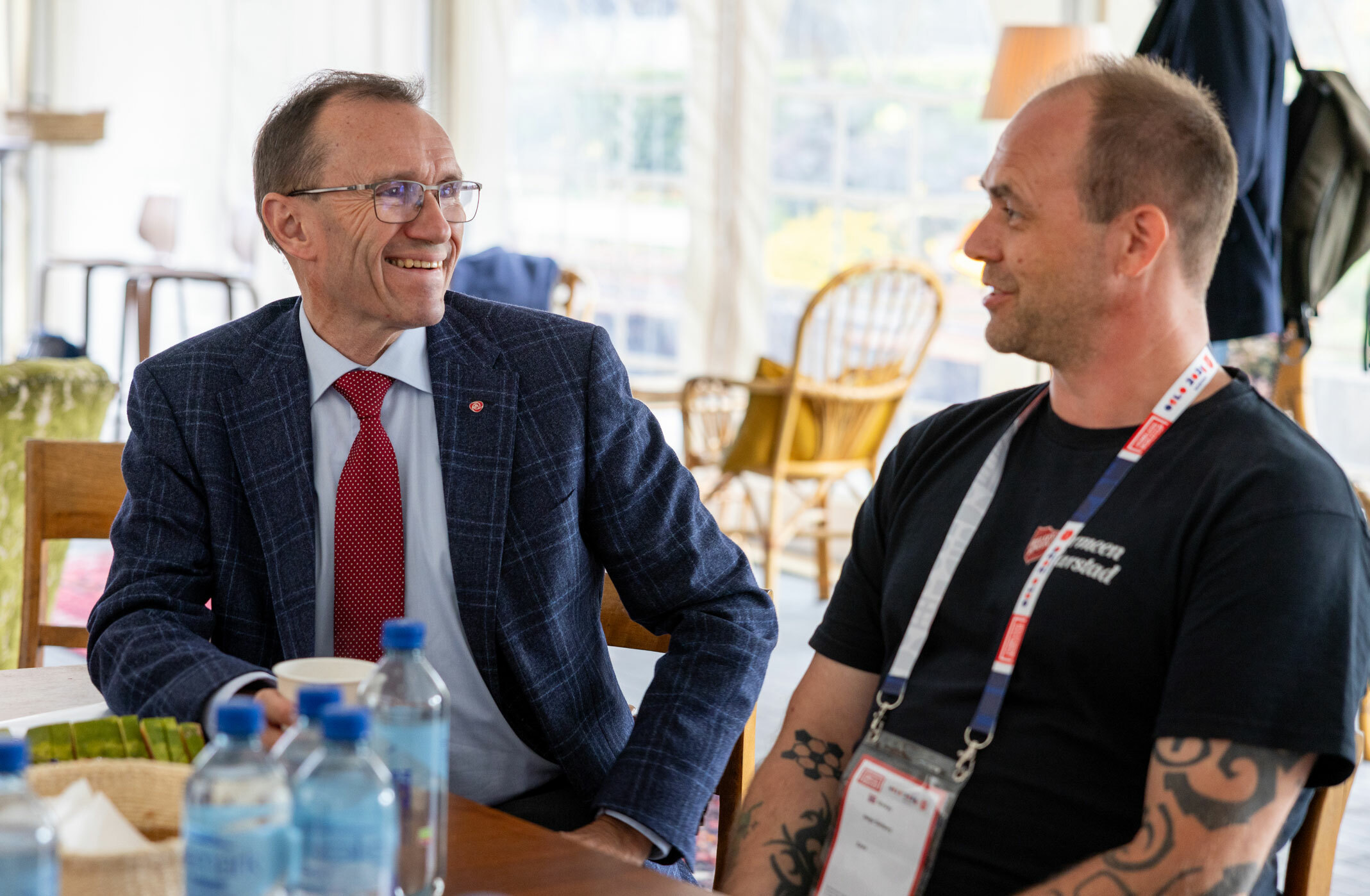 Foto: Mann med briller i mørk dress og rødt slips snakker til en mann i T-skjorte med tatoveringer. De sitter inne i et telt.