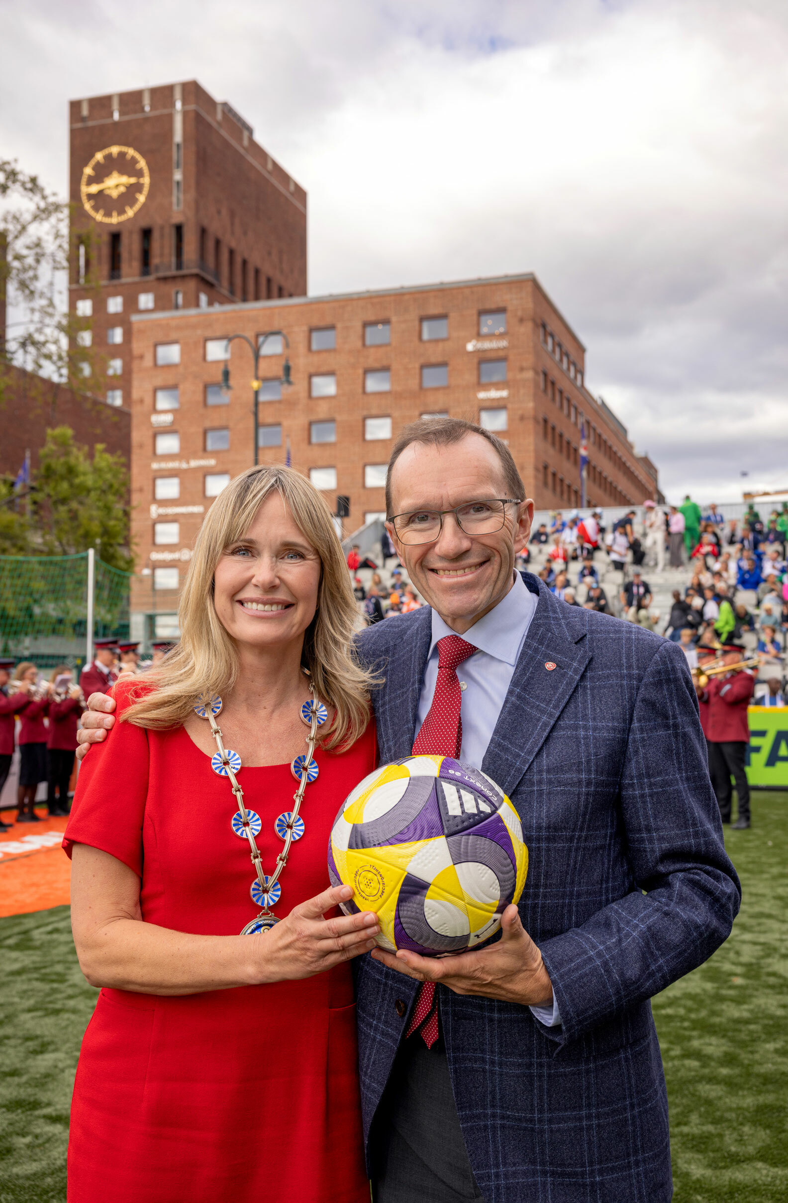 Foto: Blond kvinne i rød kjole med ordførerkjede står og holder en fotball sammen med en mann med briller i mrøk dress og rødt slips.