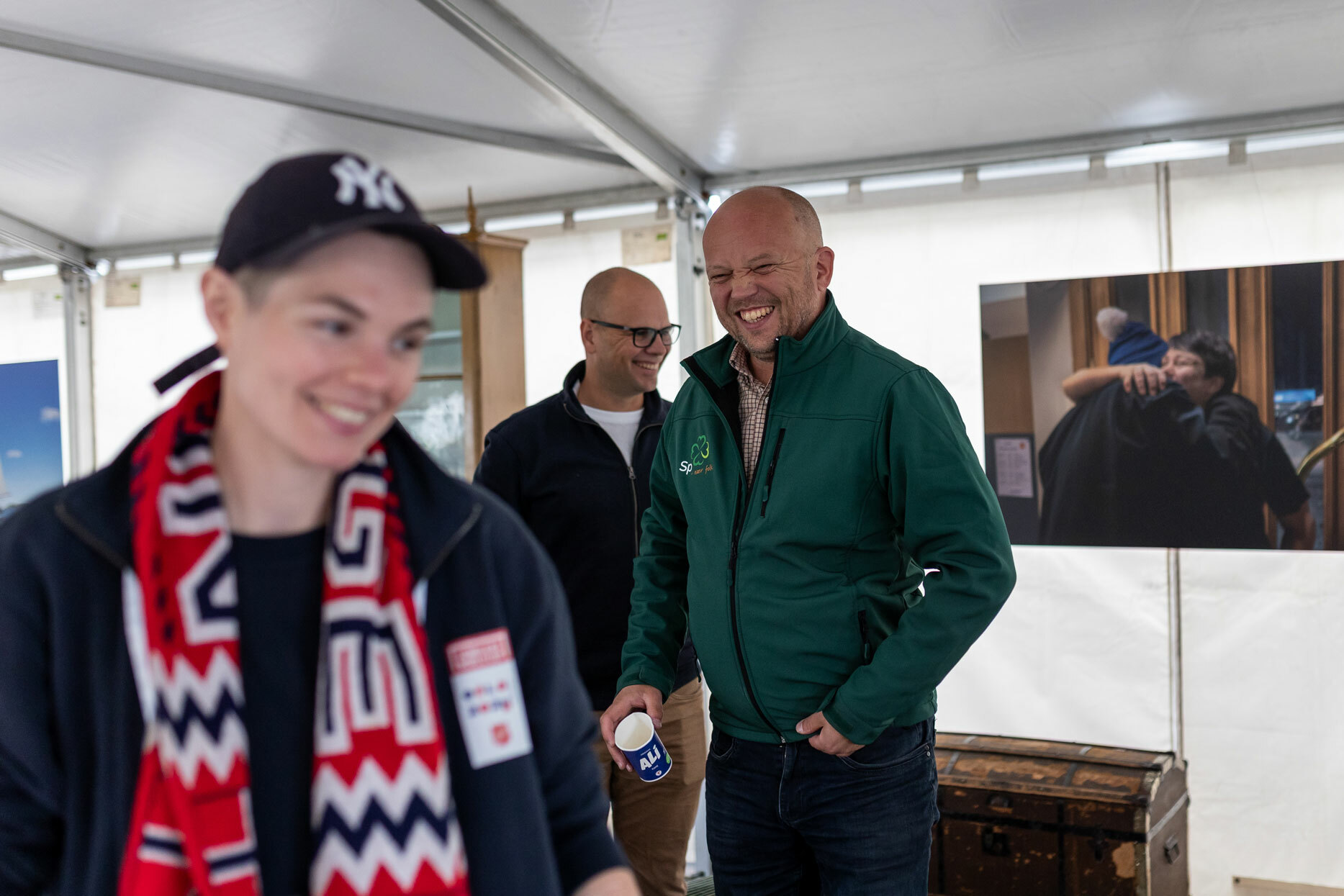 Foto: Kvinne med kort hår, caps og fotballskjerf i rødt, hvitt og blått der det står Norge. Bak står to menn med glattbarberte hoder – en med briller og en uten.