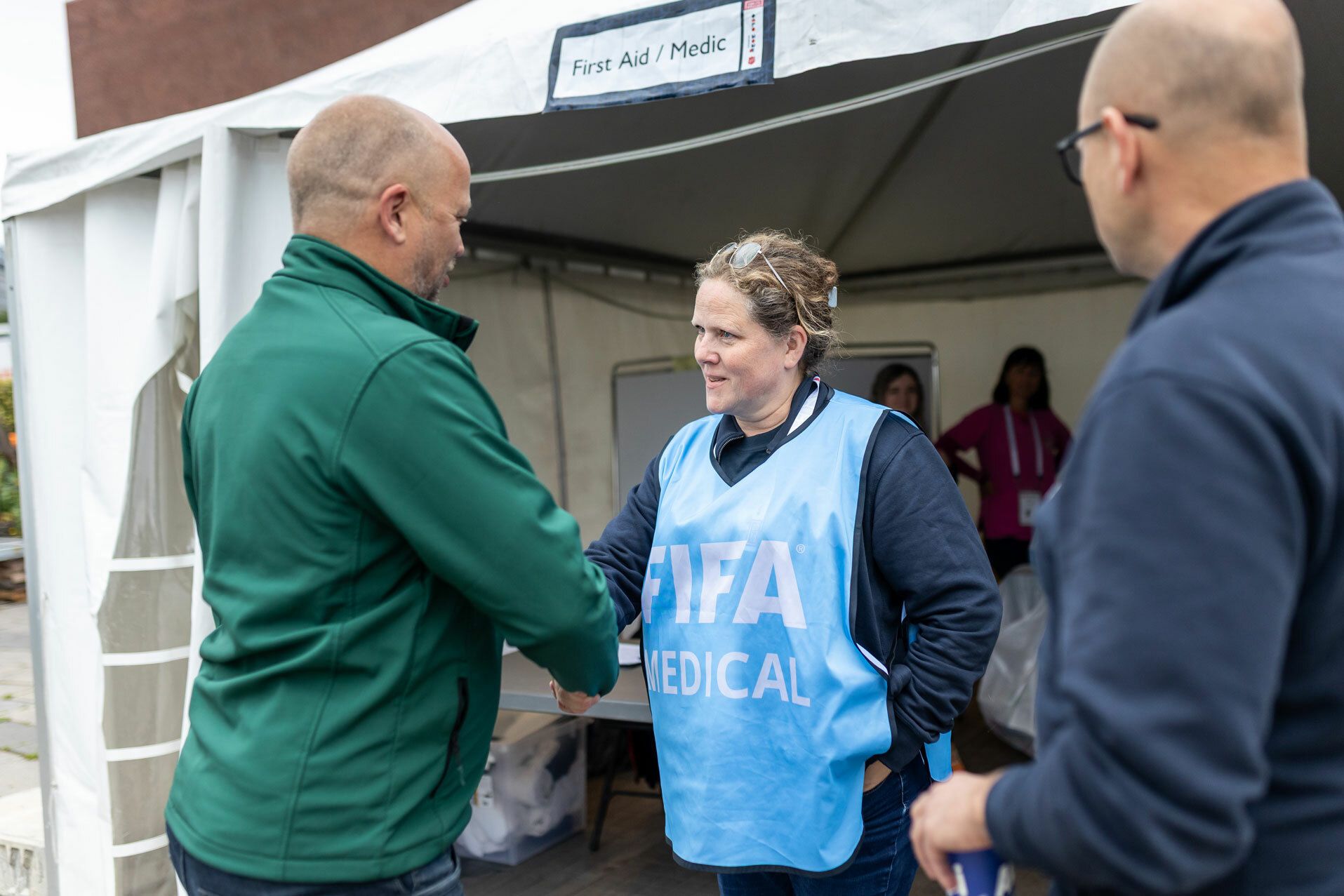 Foto: Mann i grønn jakke med barbert hode hilser på damen med Blå i åpningen til et telt der det står "First Aid / Medic" over. Ved siden av står en mann i blå jakke og briller.