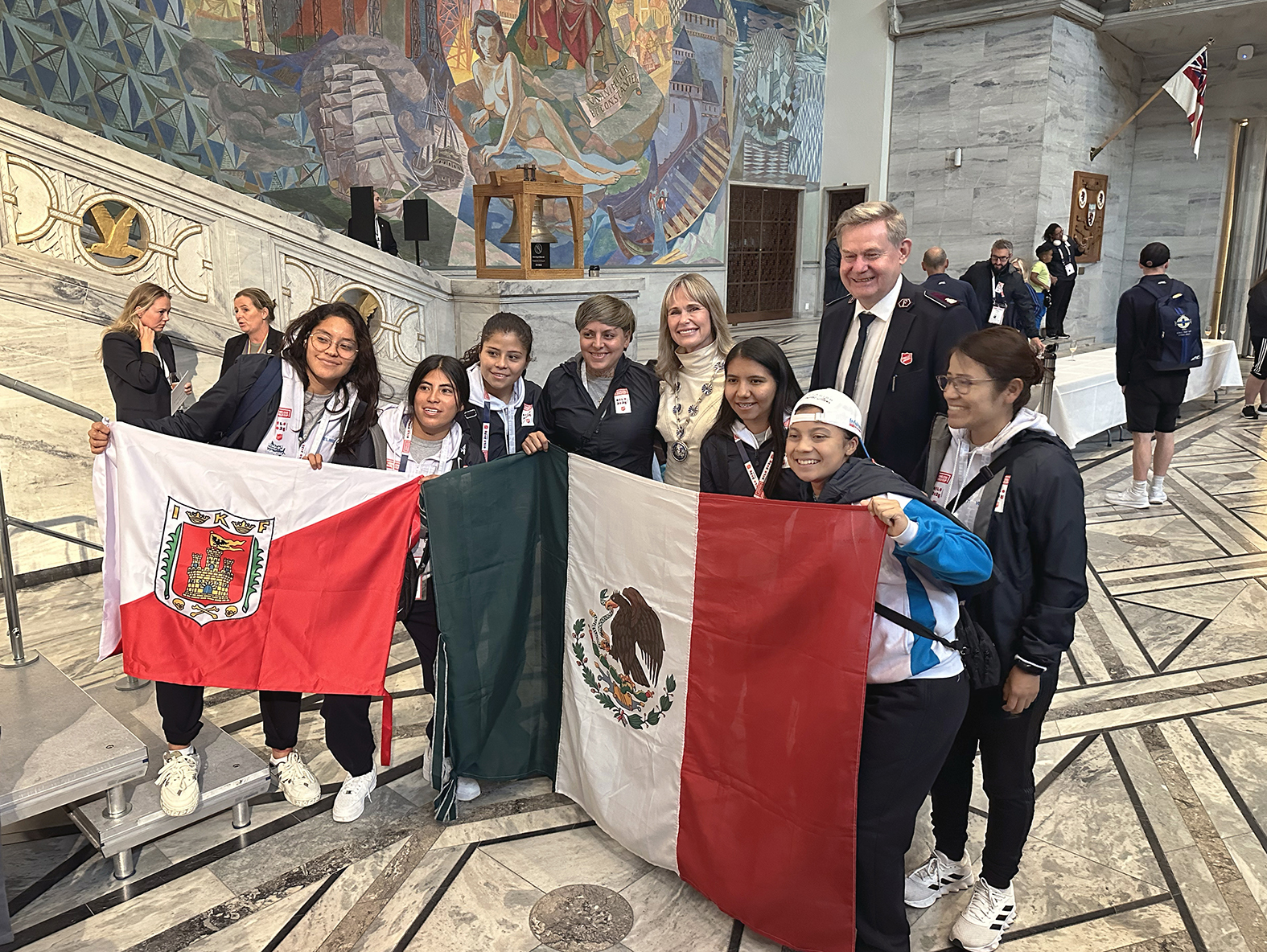 Foto: Kvinnelaget fra Mexico holder opp to nasjonale flagg. De tar bilde av seg med blond kvinne i hvit kjole med ordførerkjede og en høy mann i uniform fra Frelsesarmeen.