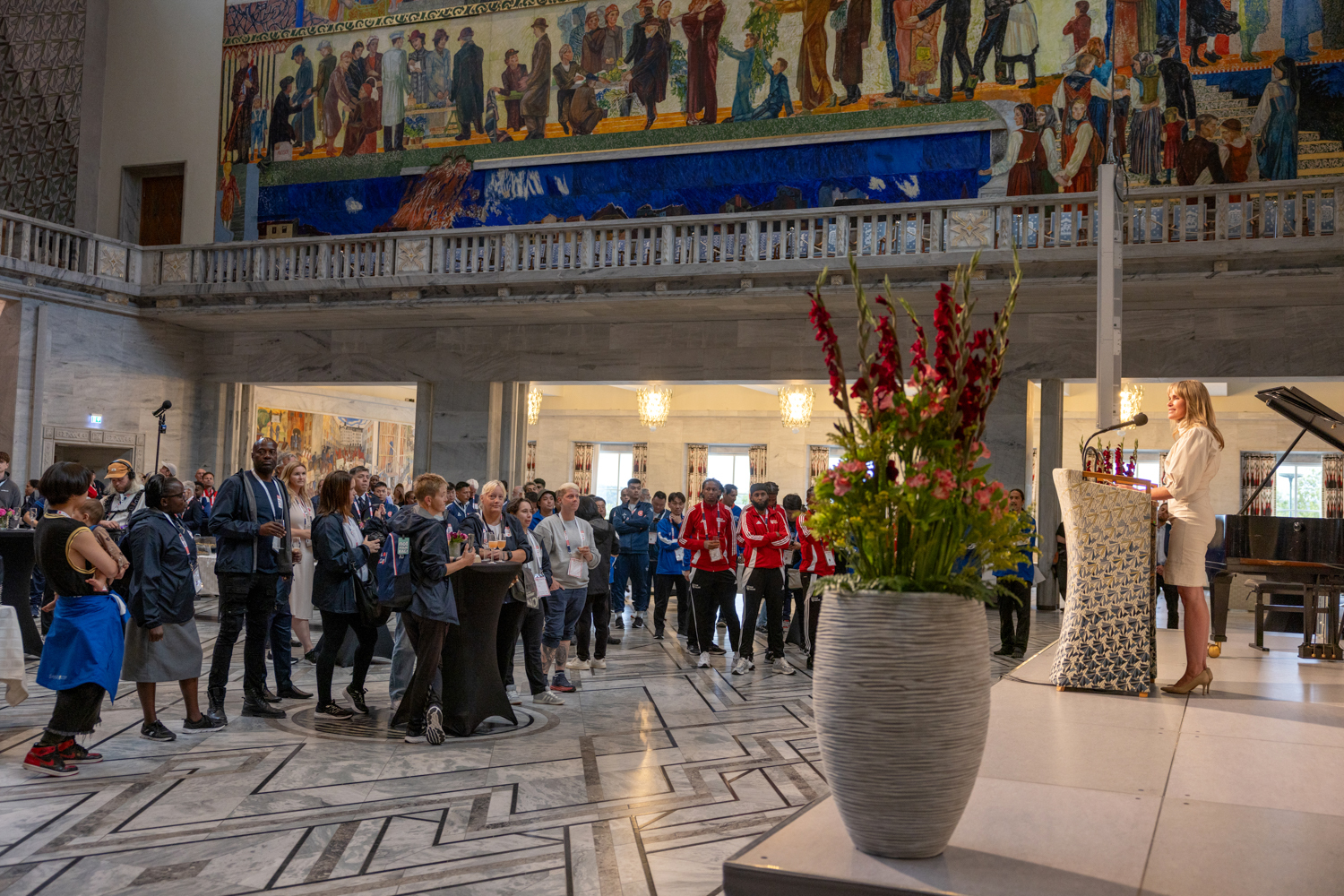 Foto: På talerstolen står ordføreren i hvit kjole og taler til folk i salen. Det er malerier på vegger og marmorgulv. Bildet er tatt fra siden, slik at kvinnen på talerstolen med blondt hår sees i profil.