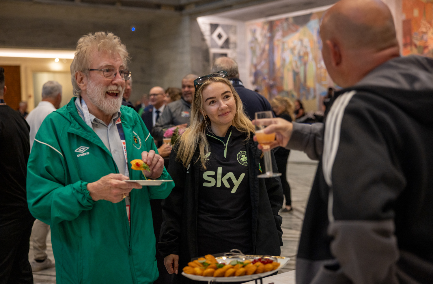 Foto: Eldre mann i grønn treningsjakke, grått hår og briller står ved siden av en ung kvinne med langt blondt hår i en hettegener der det står "sky" på brystet. De spiser kanapeer og snakker med en annen voksen mann.