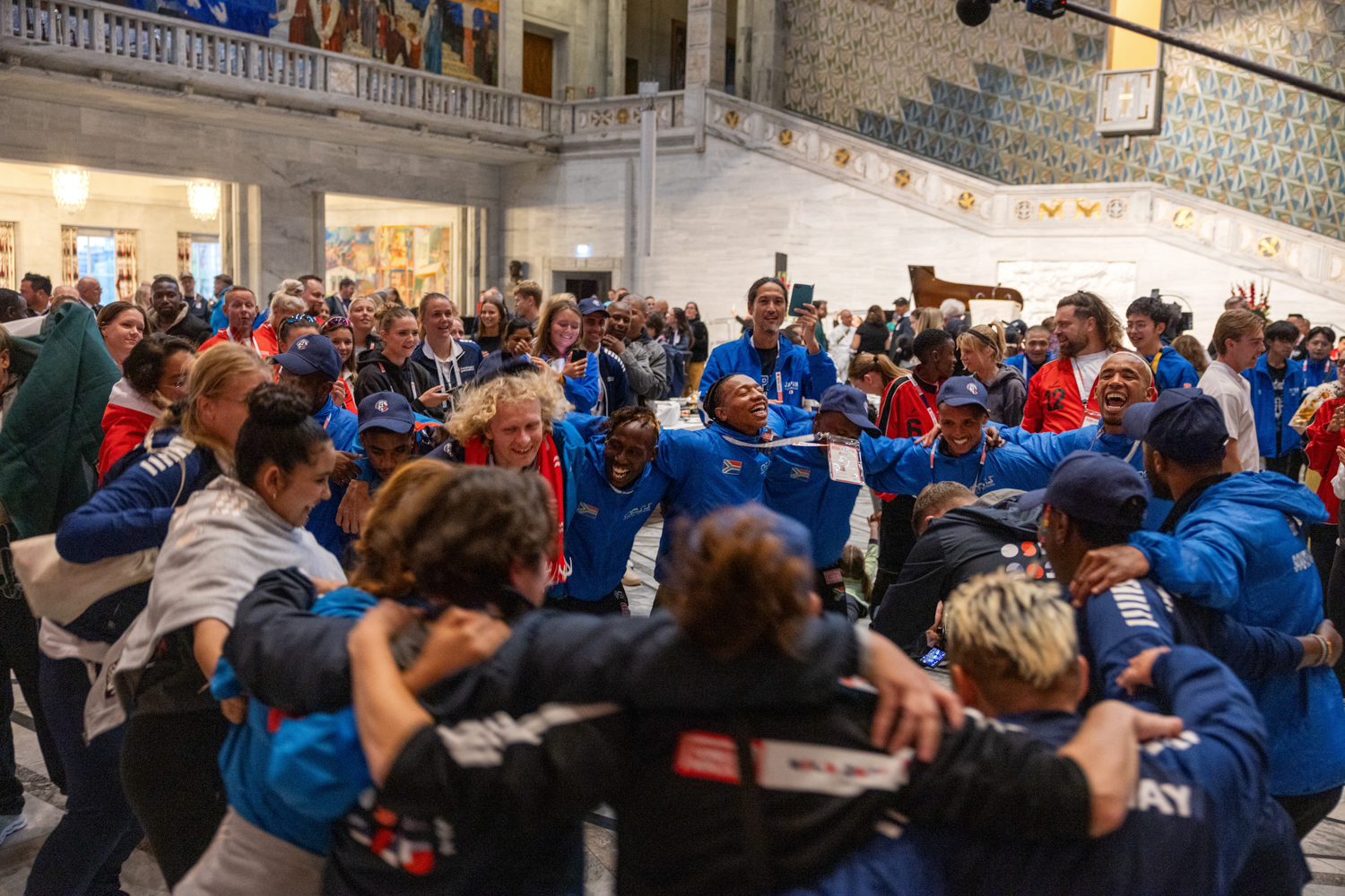 Foto: Mange folk i fotballdrakter danser i ring i inne i den ærverdige store salen i Oslo rådhus.