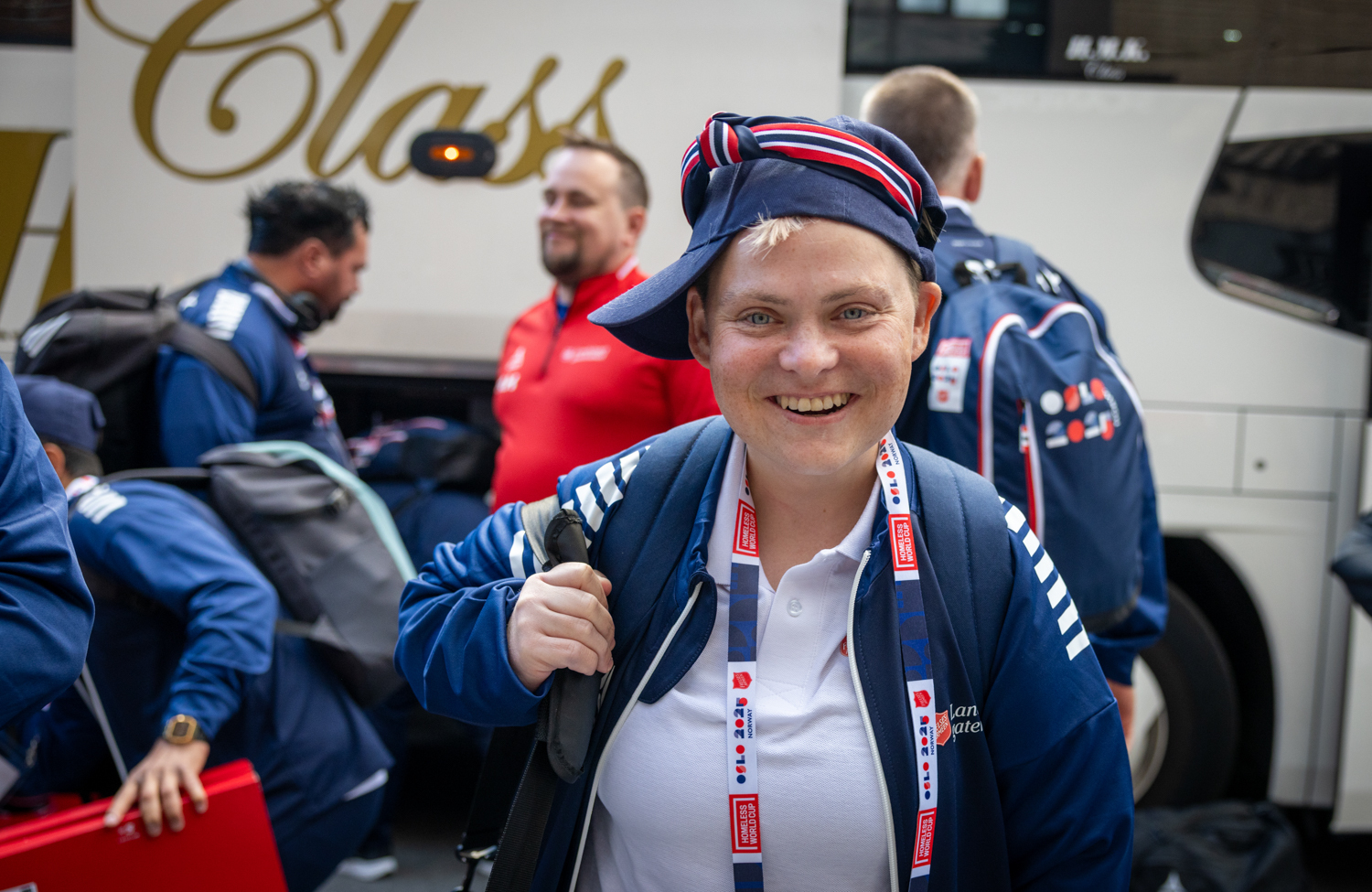 Foto: Smilende norsk jente med capsen på snei og norske farger og flagg bærer en ryggsekk og er på vei bort fra bussen.