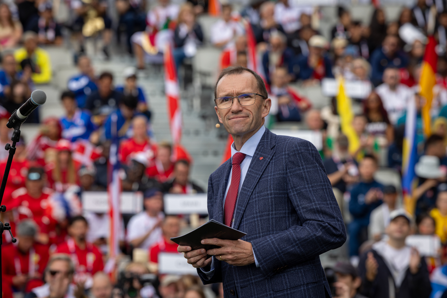 Foto: Mann med mørk hår og briller står i dress på en stadion med fulle tribuner.