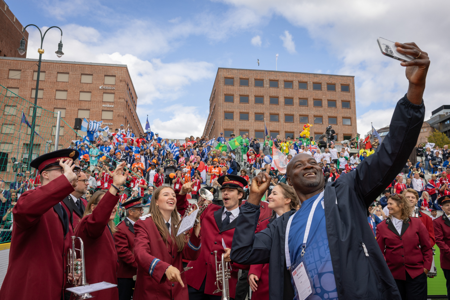 Foto: Svart mann tar selfie sammen med musikanter i uniform og fullstappet tribune bak.