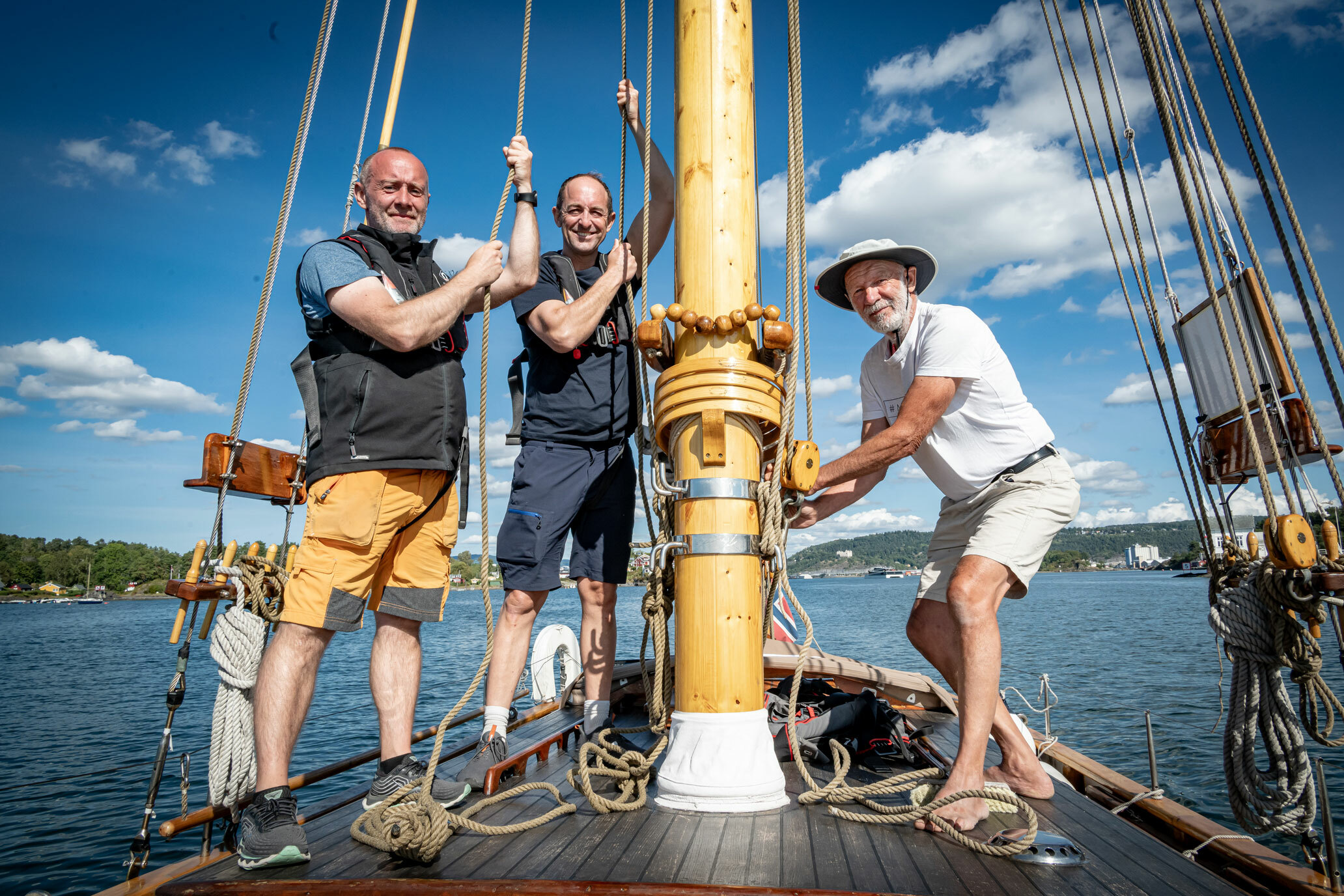 Foto: Tre menn står ved en mast og drar i tau. Poserer litt for fotografen.