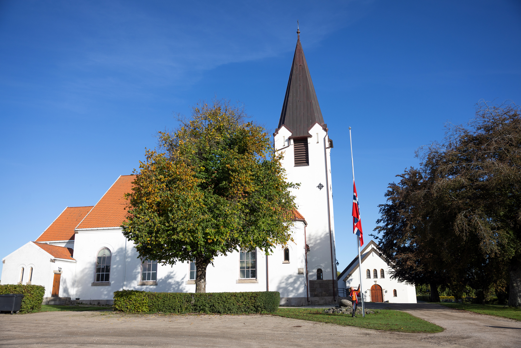 Hvit kirke med flagget på halv stang utenfor. Store trær Blå himmel bak.