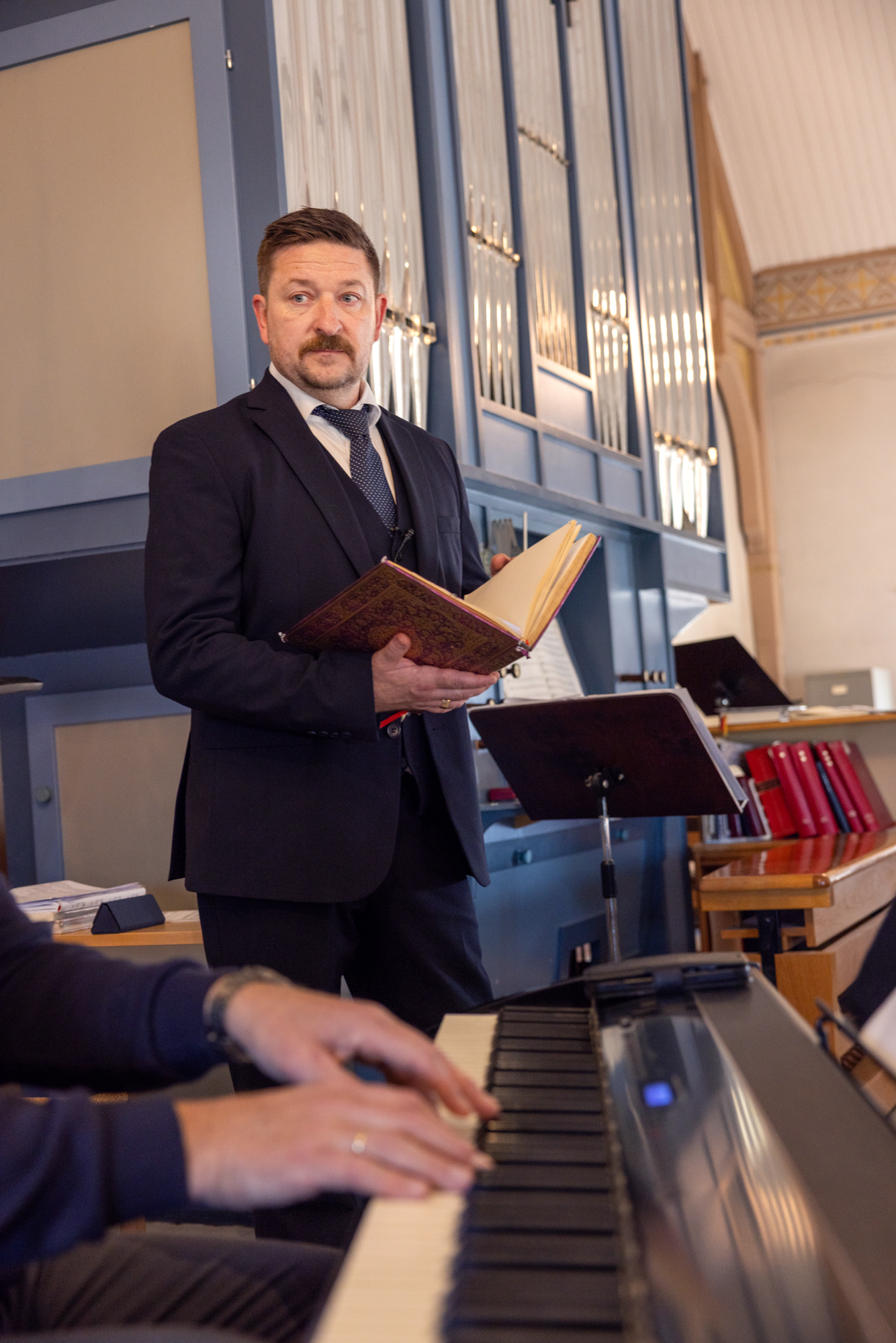 Mann står med en oppslått sangbok foran et orgel. I forgrunnen ser vi hender som spiller på et piani.