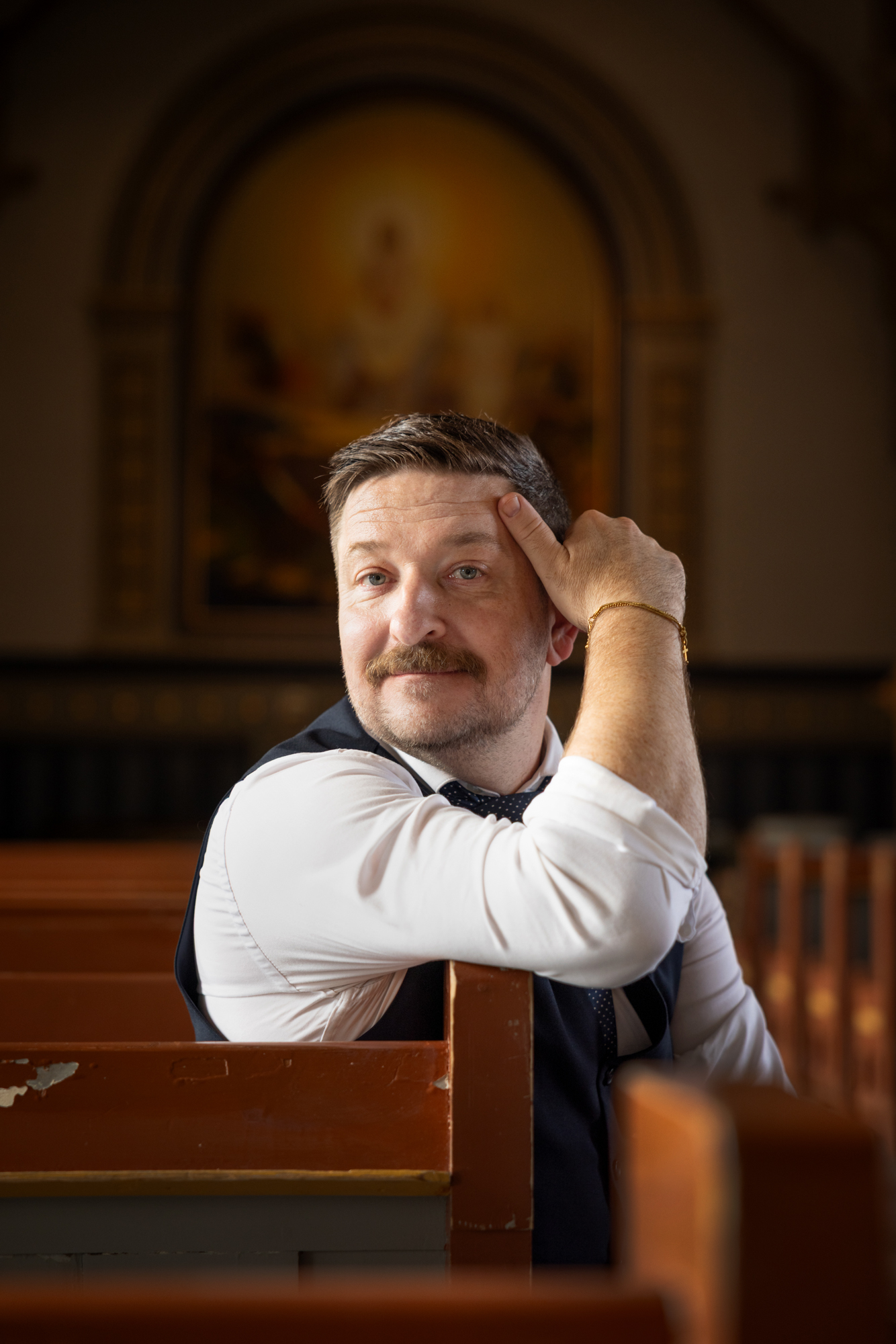 Mann med mørkt hår og bart sitter på en kirkebenk med ene armen opp mot hodet. Han har en gullenke rundt høyre håndledd. I bakgrunnen er det en religiøs utsmykning i form av et maleri av en oppstanden Jesus.