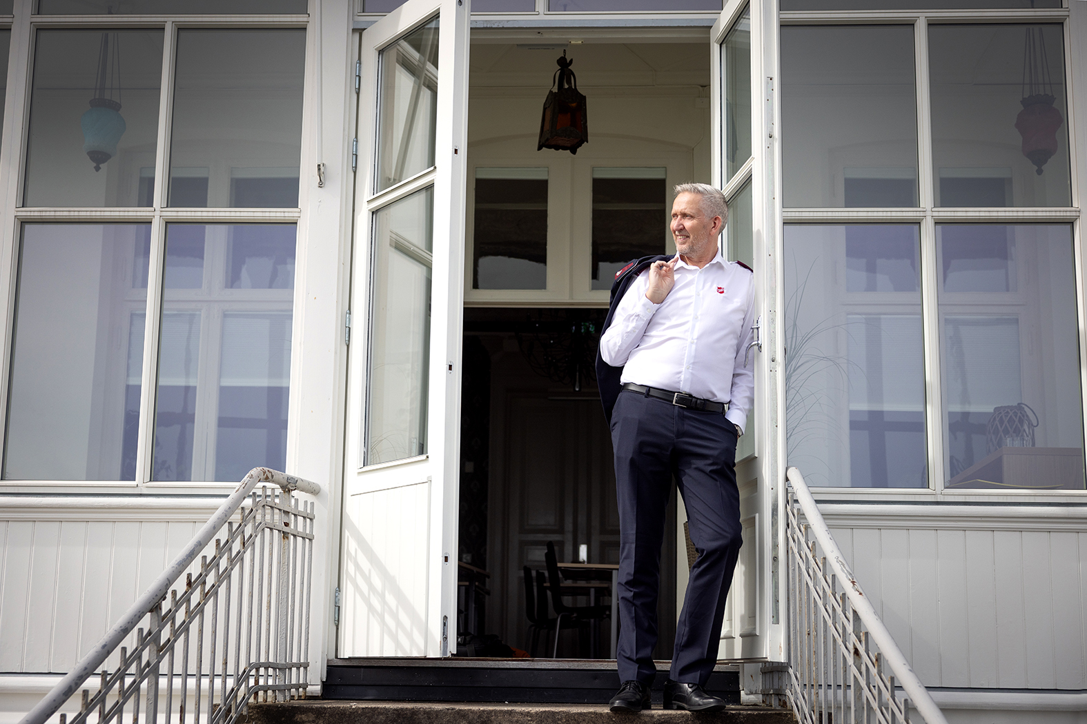 Foto: Mann i hvit skjorte og mørke dressbukser står på øverste trappetrinn foran to åpne dører til et gammelt hus. Han har jakken hengende over skulderen.