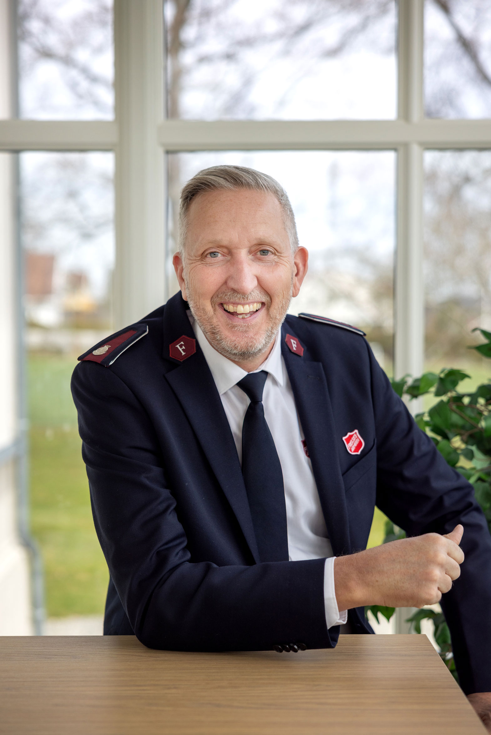 Foto: Mann i uniform fra Frelsesarmeensitter ved et bord. Han er tidlig i 60-årene og smiler bredt. Gjennom den hvite glassdøren bak ham ser vi en grønn plen-