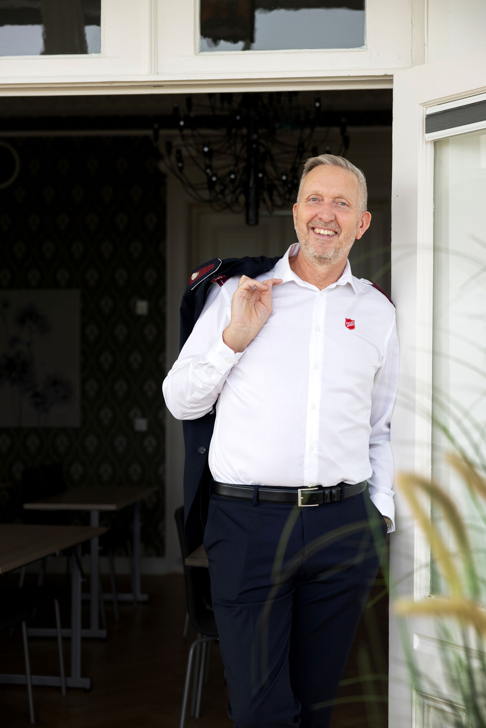 Foto: Mann med kort, grått hår står i en døråpning. Han har på seg hvit skjorte og holder uniformsjakken liksom bak på ryggen slik vi ser i Dressman-reklamer.