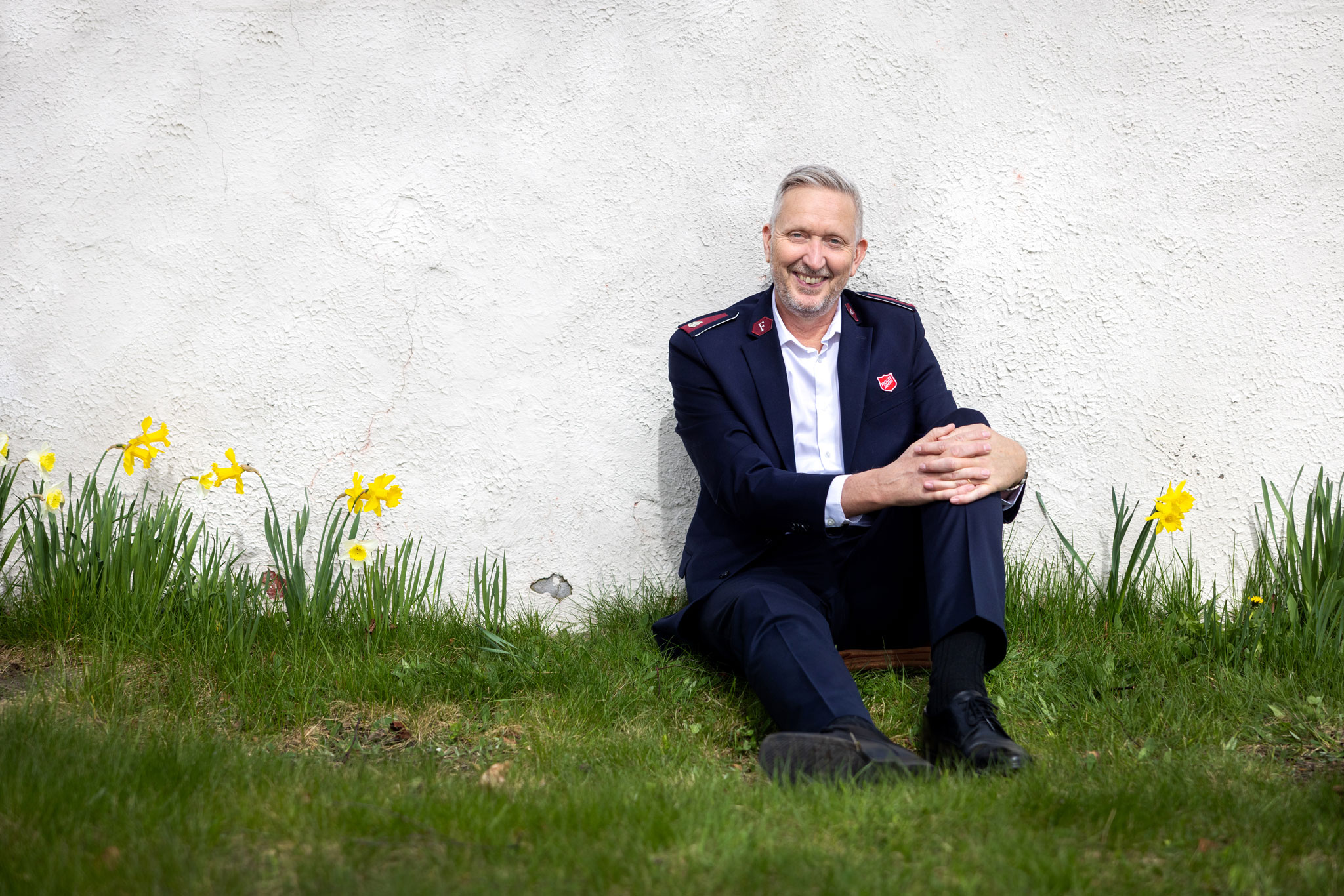Foto: Mann tidlig i 60-årene sitter inntil en hvit murvegg. På den grønne plenen står det gule påskeliljer. Han har på seg en mørk uniform fra Frelsesarmeen og hvit skjorte.