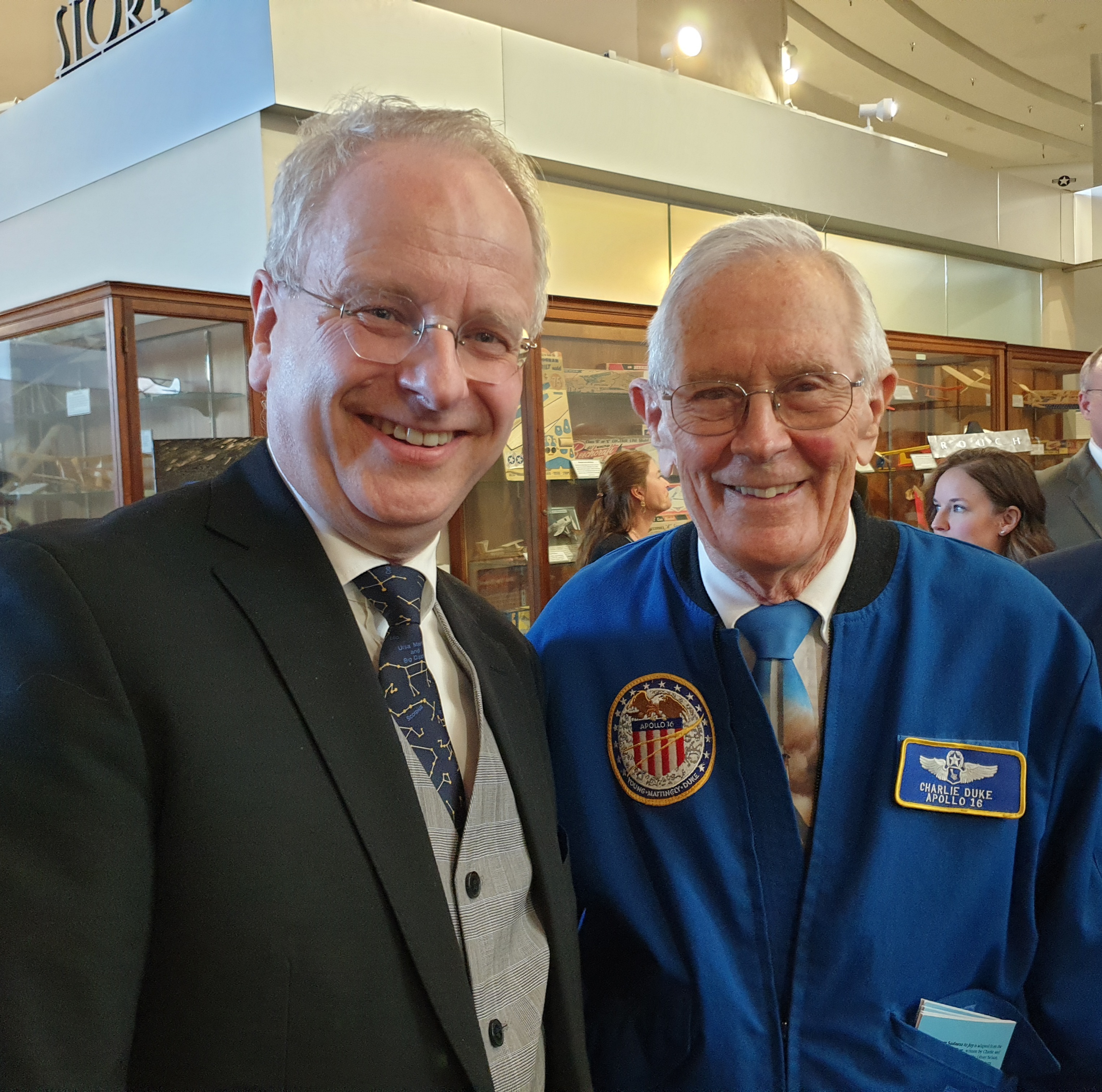 Foto: En norsk mann i slutten av 50-årene står sammen med en pensjonist i en knall blå jakke med merke fra Apollo 16 og et annet pilotmerke. Begge har briller og grått hår.