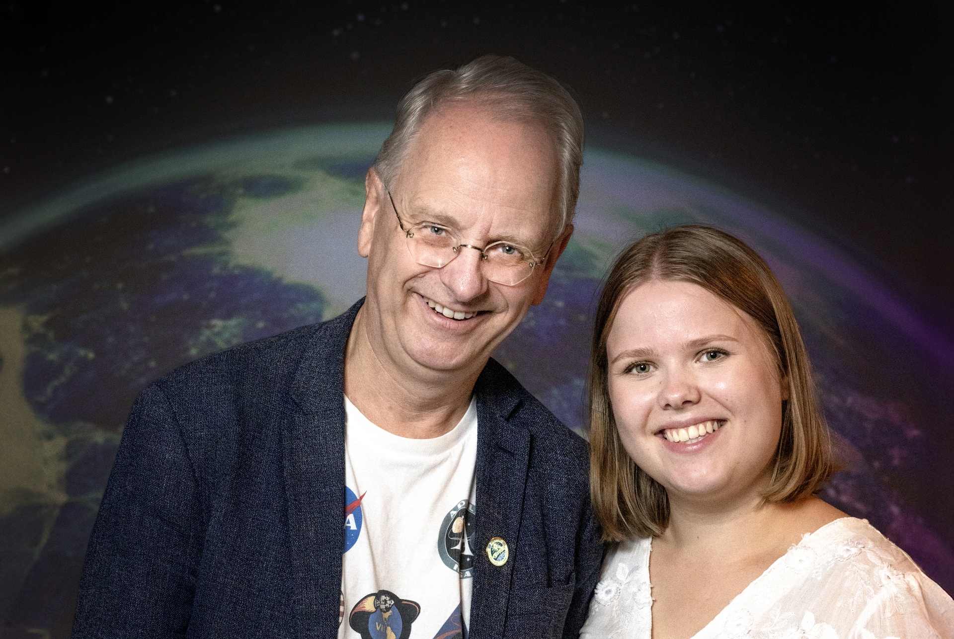 Foto: Mann i slutten av 50-årene sammen med ung kvinne i 20-årene. Han er gråhåret og har briller hun er mellomblond. De står foran et bilde av vår klode.