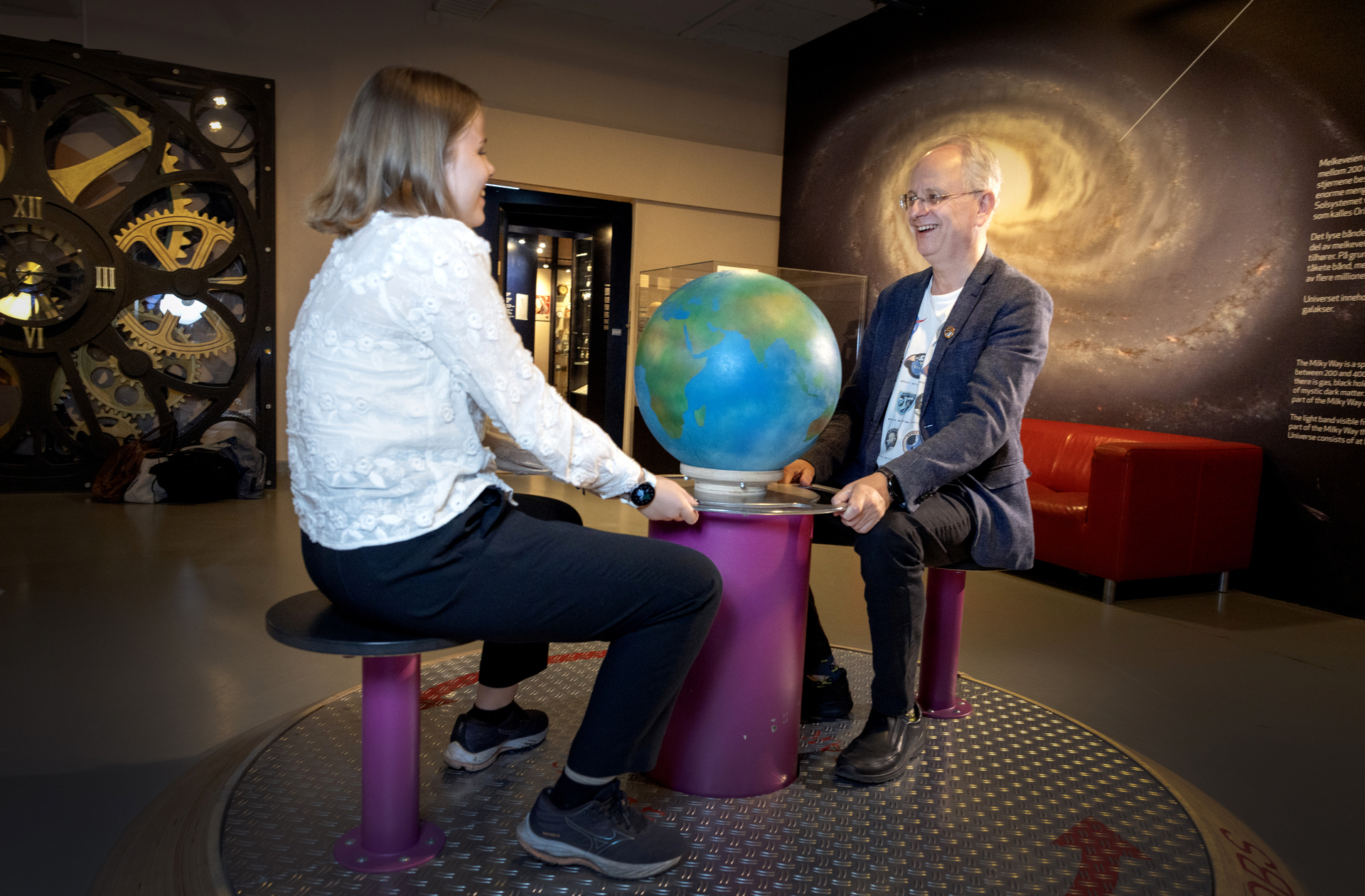 Foto: Voksen mann med briller og dressjakke sitter rett mot en ung jente i hvit bluse. De sitter på hver sin krakk som er festet til et lite bord med en globus på.