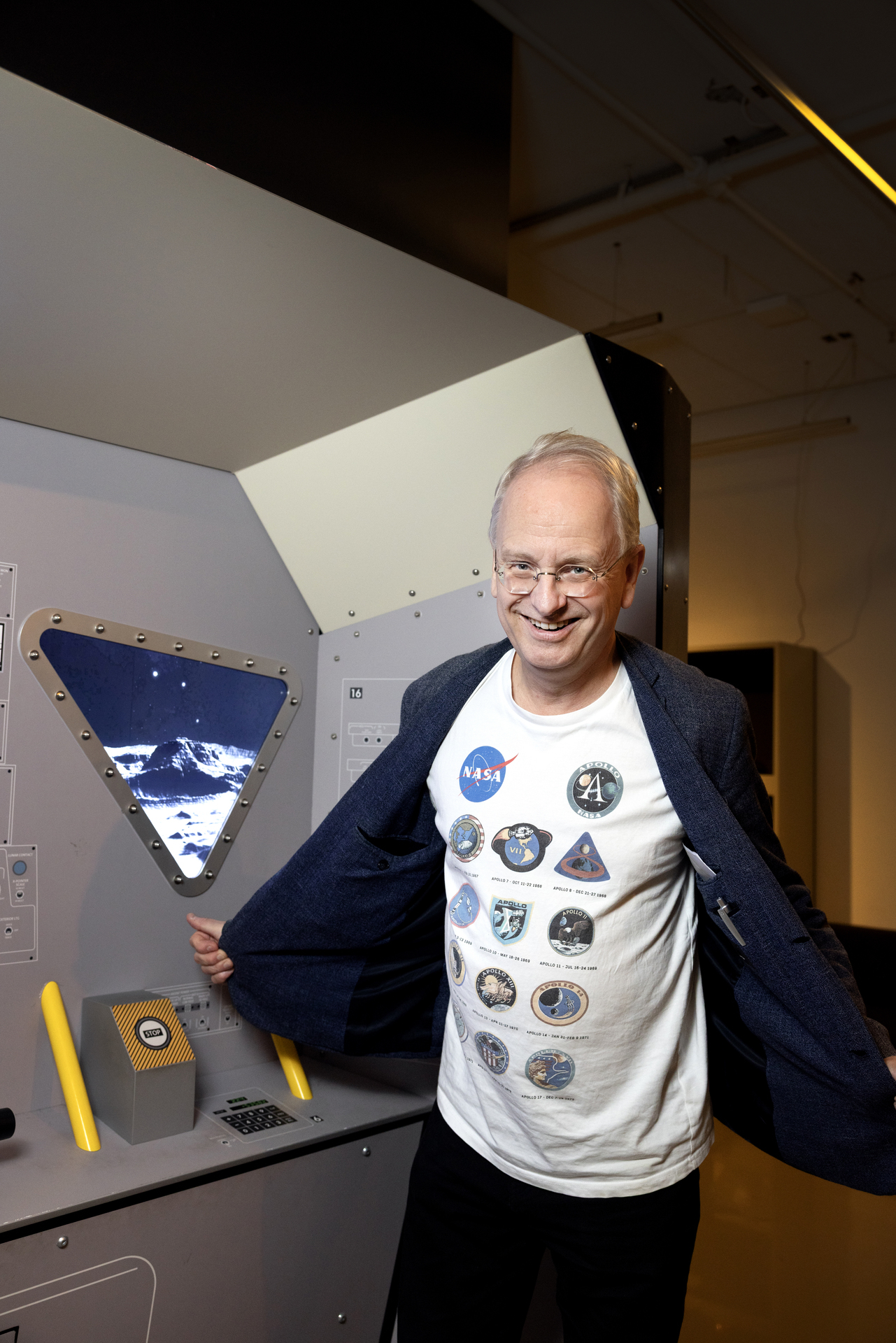 Foto: Mann med briller åpner den blå dressjakken og viser fram den hvite T-skjorten sin med flere symboler.