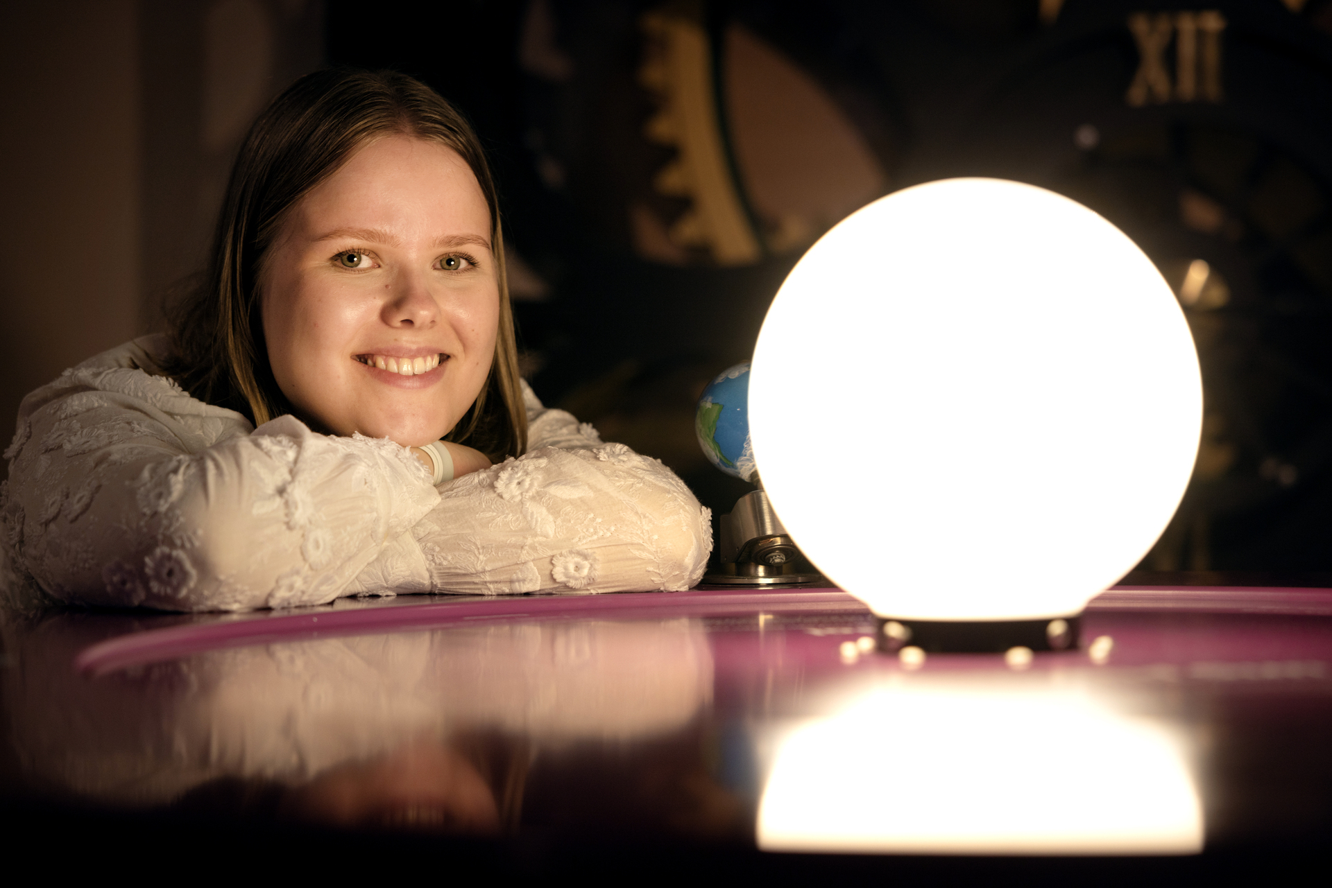 Foto: Ung kvinne ligger med begge armene på et bord med en stor, gul lampe midt på som skal forestille solen.