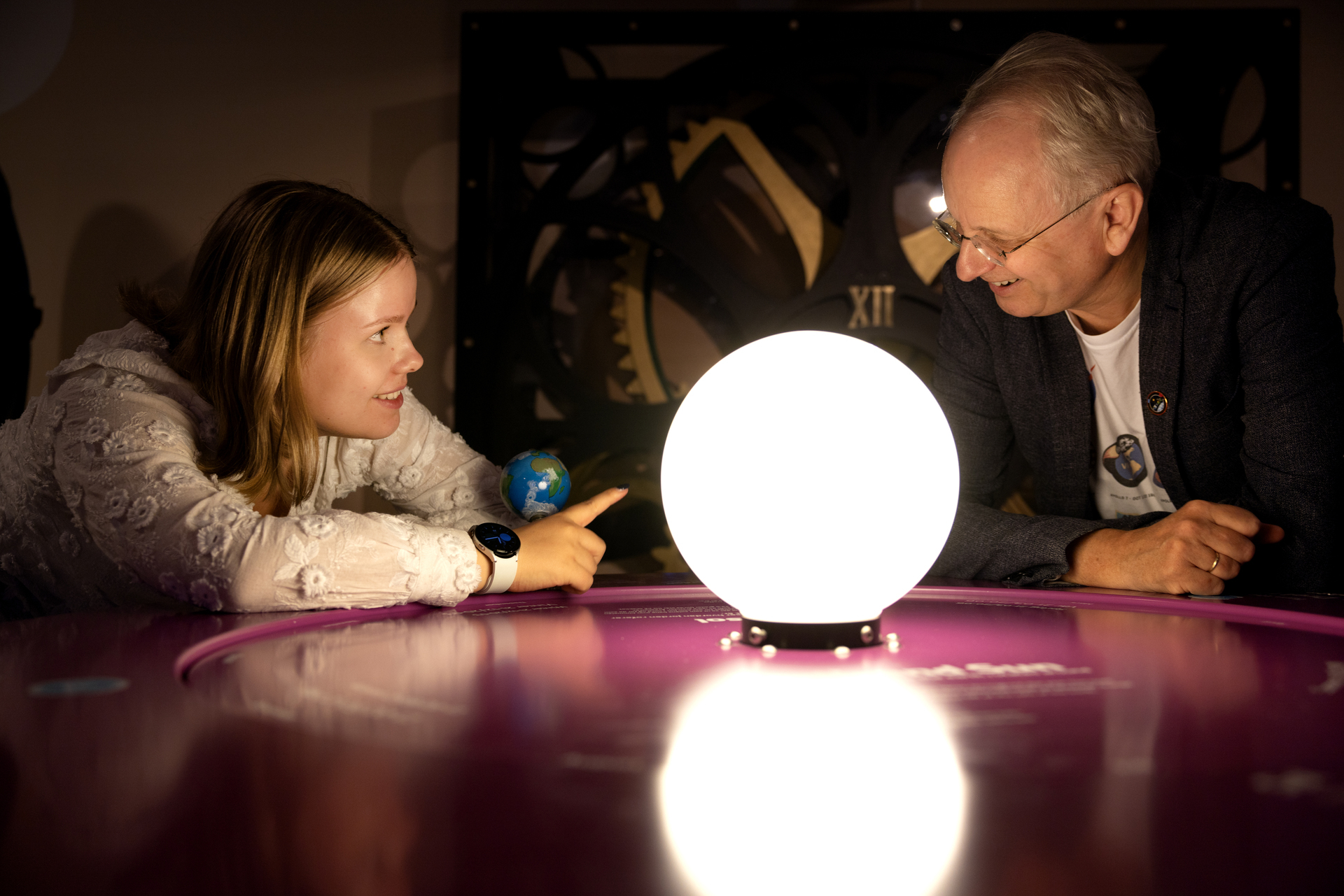 Foto: Rundt den samme lampen (solen) på bordet peker den unge kvinnen mot en mann med briller.