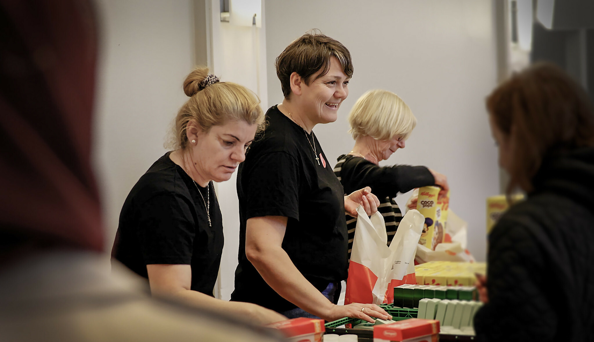 Tre kvinner fra Frelsesarmeen deler ut mat og poser til folk som kun vises som skygger i bakgrunnen.