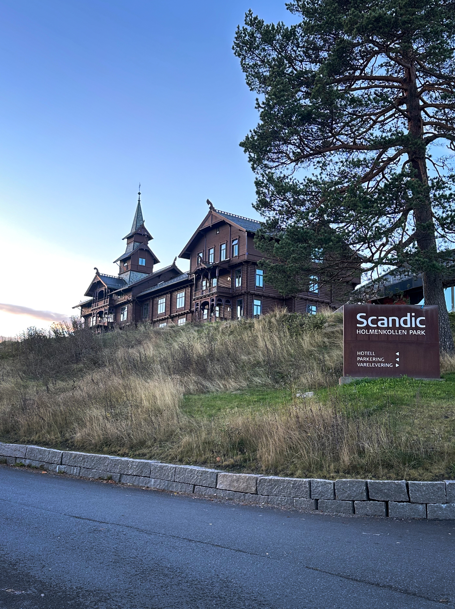 Gammelt, brunt us i dragestil i solnedgangen. På et skilt, ved en diger gran, står hotellets navn.