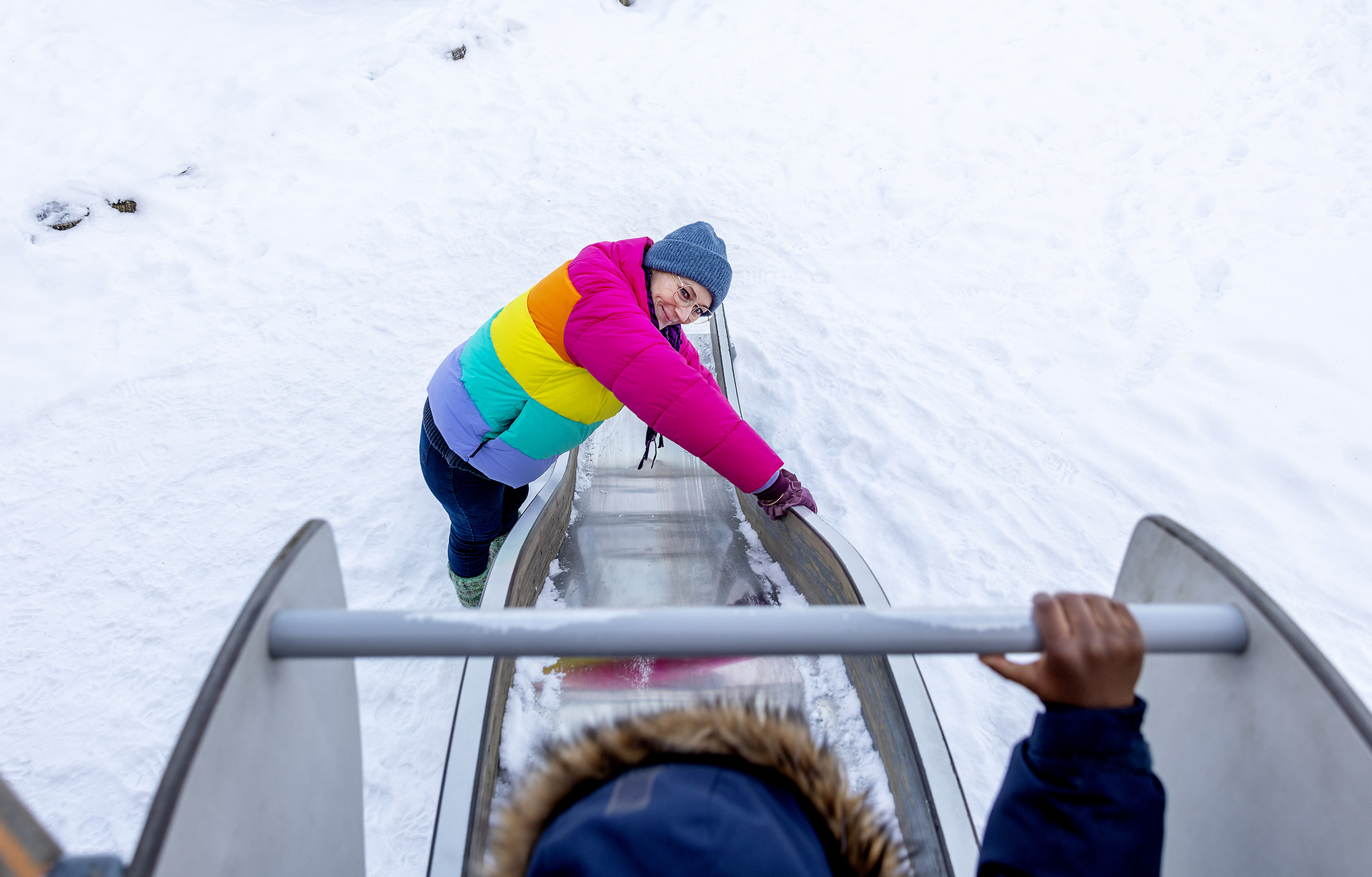 Foto: Kvinne i 50-årene står nede ved en sklie. På toppen ser vi hetta til barnet som skal sette utfor. Damen har på seg en fargerik jakke og det er snø på bakken.