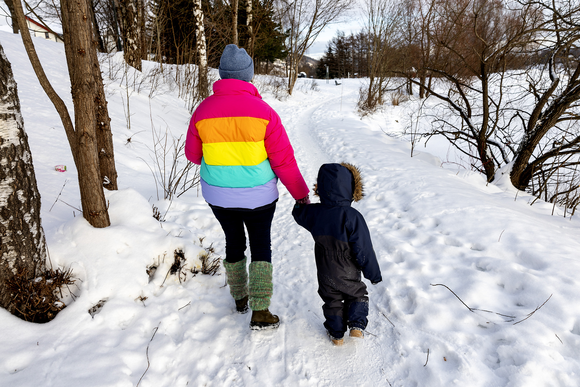 Foto: Dame i fargerik jakke leier et lite barn i blå parkdress på en sti i snøen.