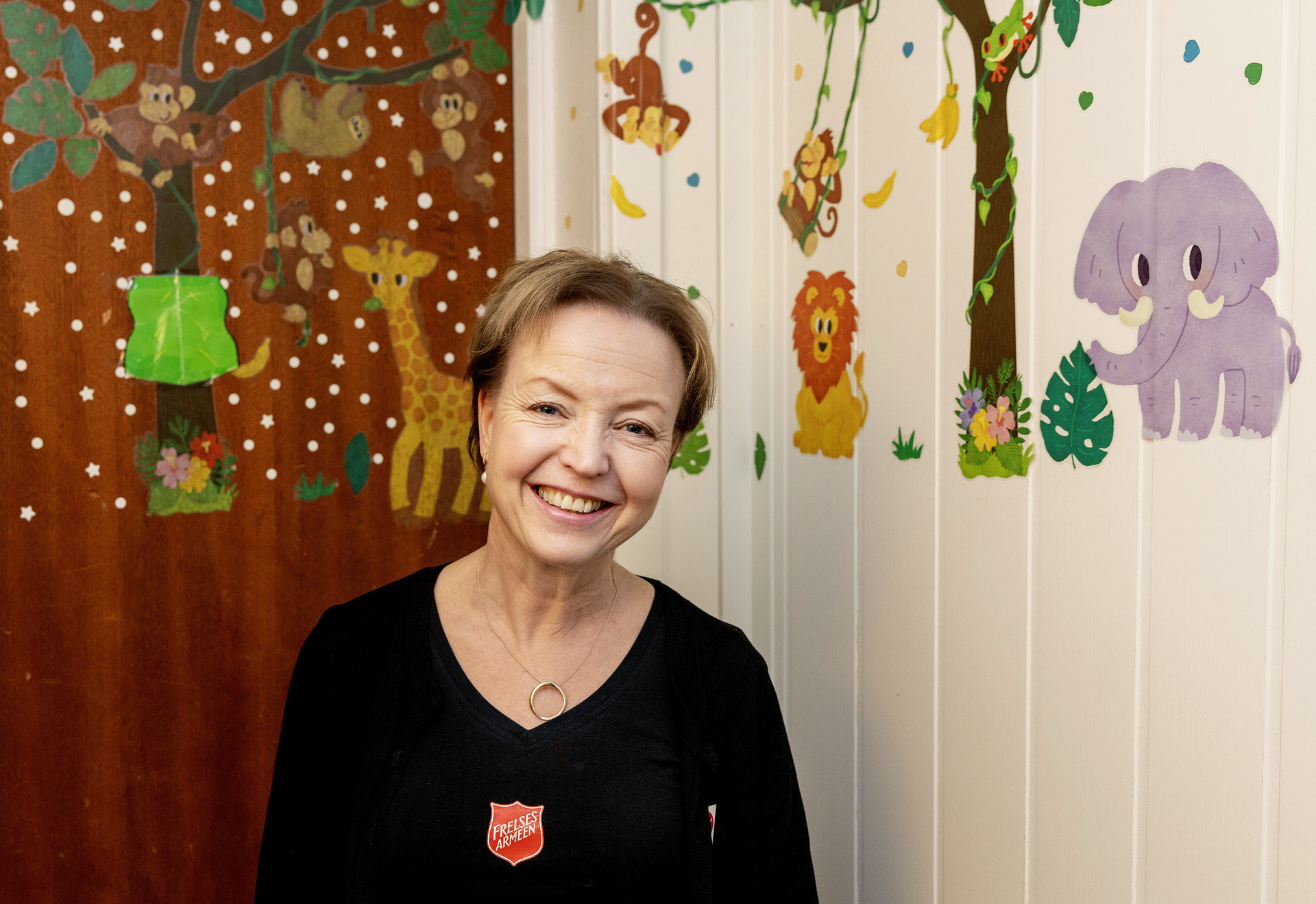 Foto: Kvinne i 50-årene med kortklippet, blondt hår smiler. Hun står i et barnerom med klistremerker av dyr fra Afrika. Hun har på seg en svart genser og en logo fra Frelsesarmeen.