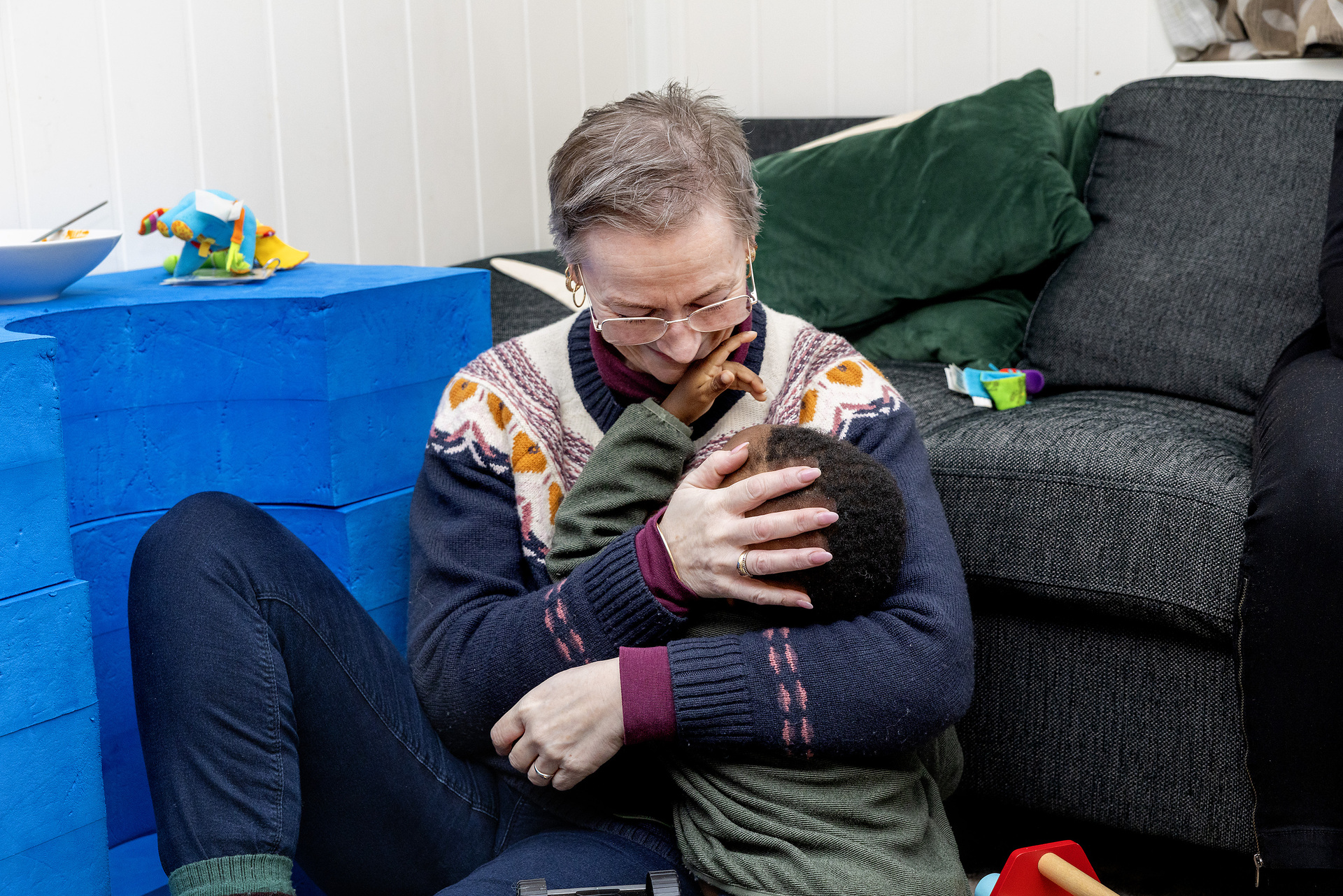Foto: Voksen dame i strikkegenser med mønster sitter på gulvet og gir en liten gutt med mørk hud og krøller en klem.