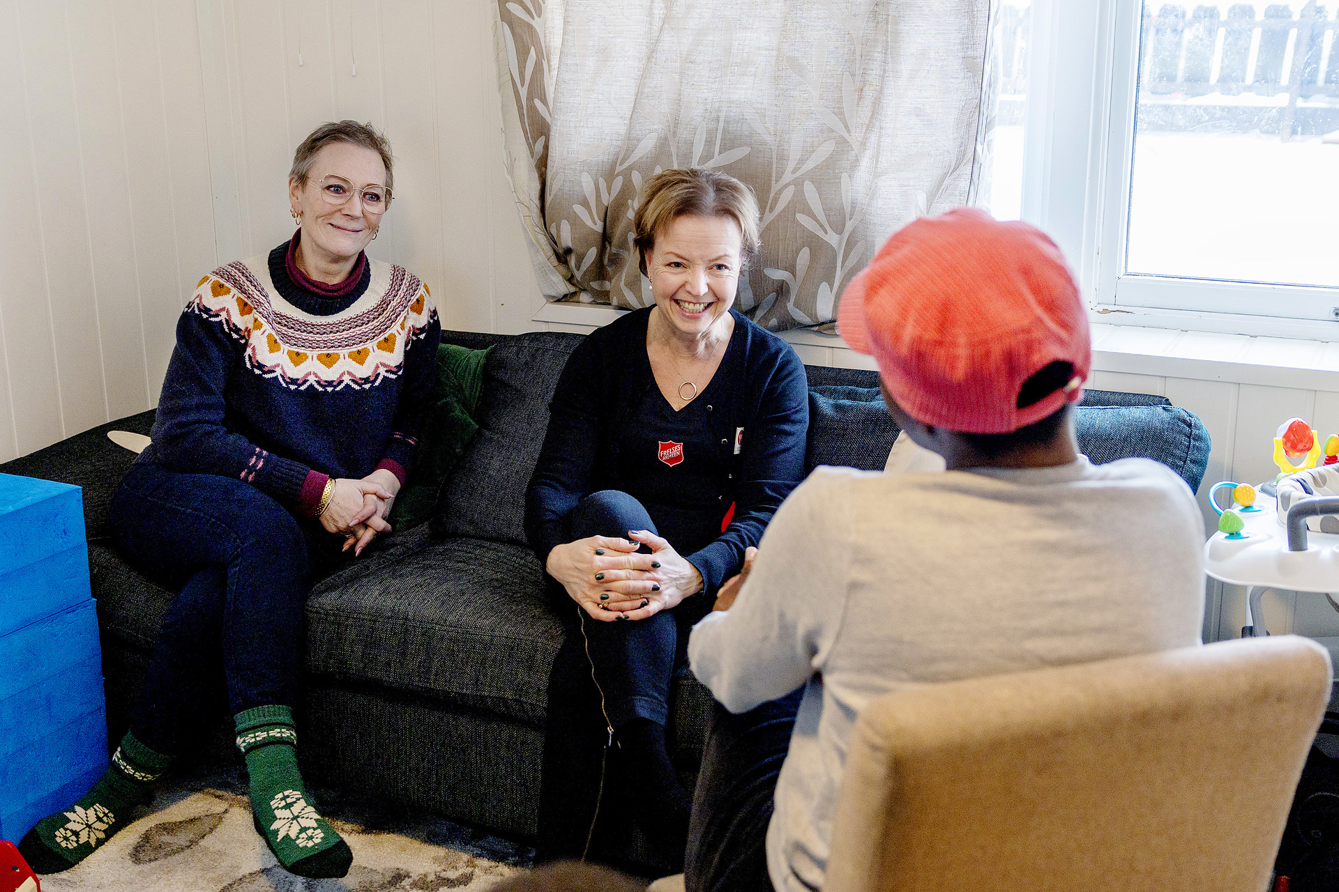 Vi er i en stue. Moren Amina ser vi bakhodet på, mens frivillig Berit sitter i sofaen til venstre med ansiktet mot kameraet, og koordinator Anne Cathrine til høyre, også  med ansiktet vendt mot kamera.