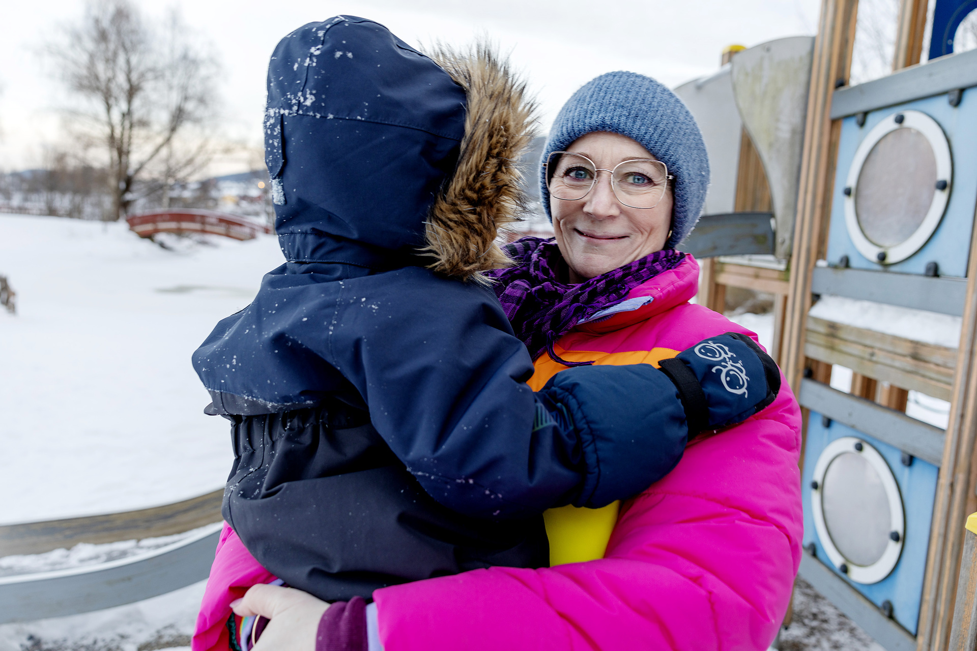 Foto: Dame i 50-årene står i en fargerik boblejakke på en lekeplass ute i snøen. I armene sine holder hun et lite barn i mørkeblå parkdress med hetten trukket over hodet.
