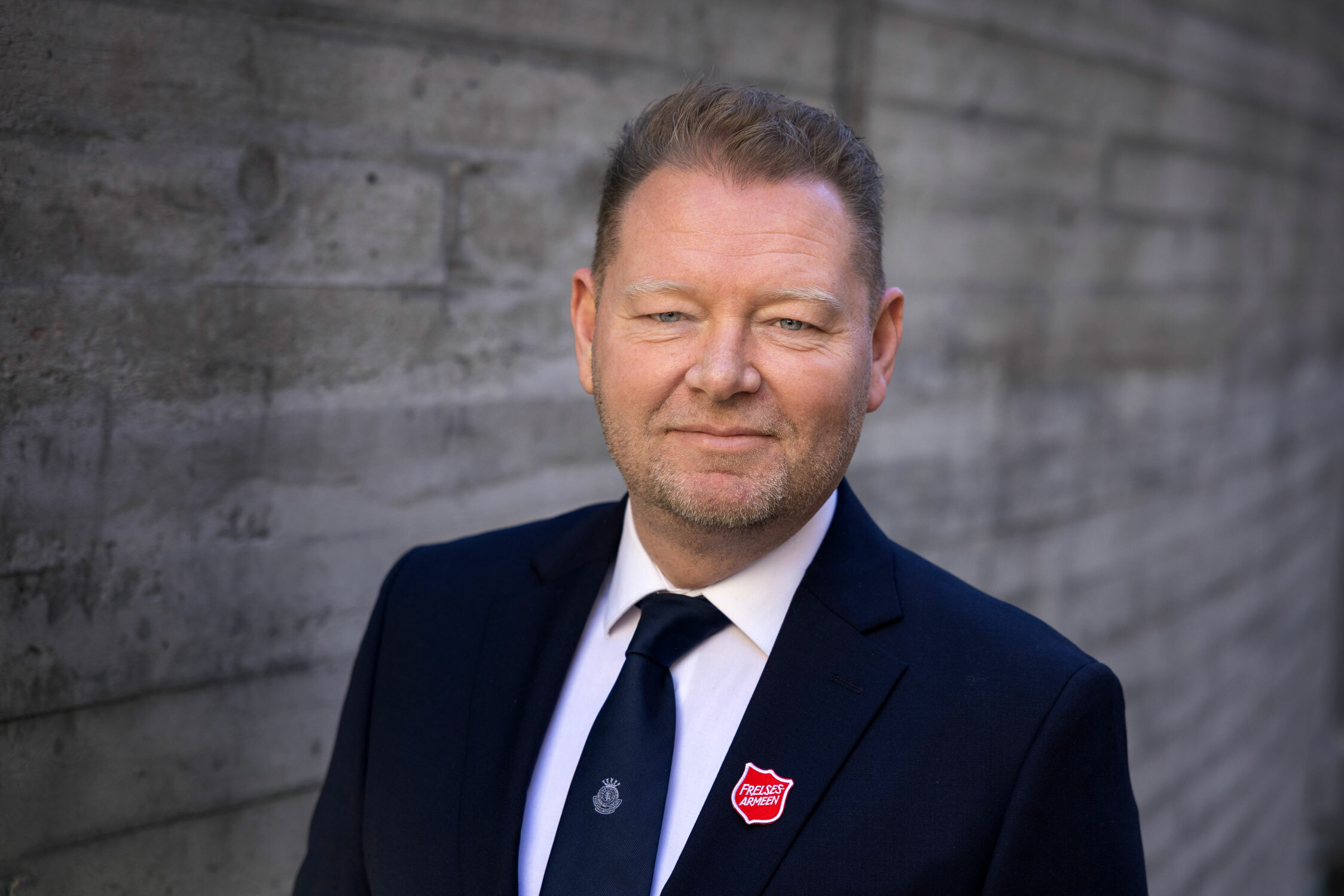 Foto: Mann i blå uniform fra Frelsesarmeen står foran en murvegg. Han er mørk blond.