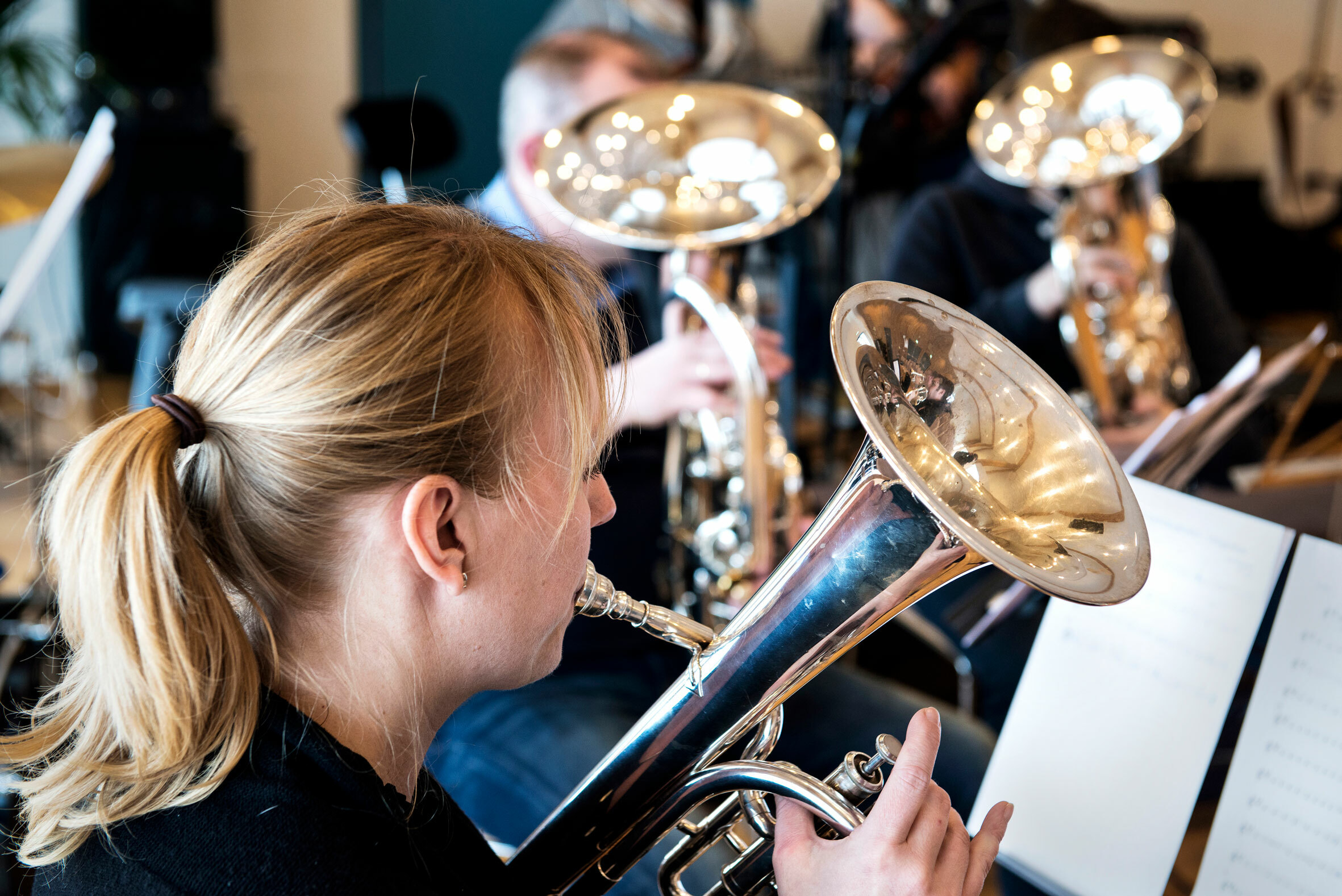 Foto: Kvinne med hestehale som spiller et mindre brassinstrument. I bakgrunnen sitter det flere og øver.