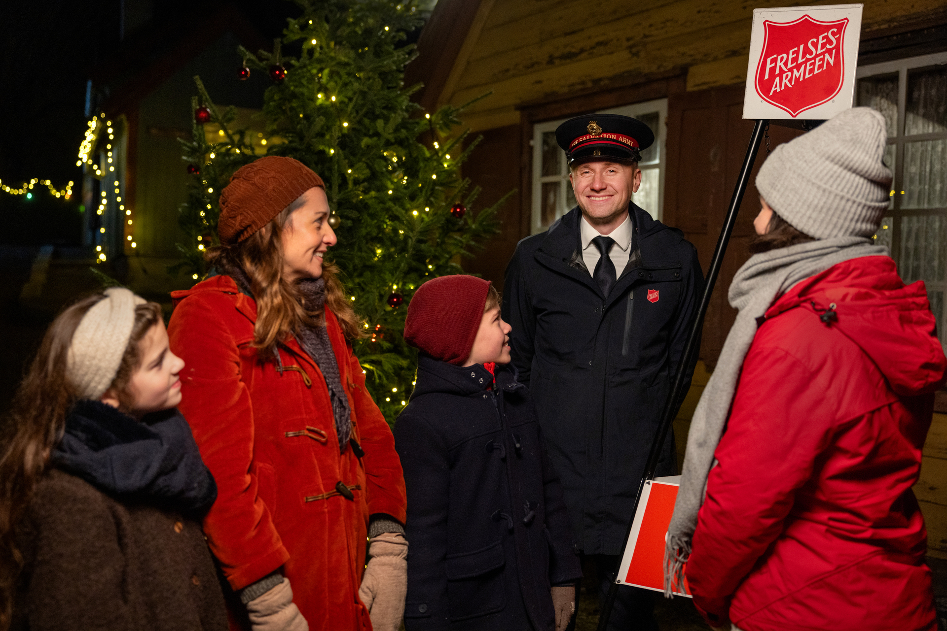 Foto: Ung mann i uniform og lue fra Frelsesarmeen står ved en julegryte. I bakgrunnen ser vi julelys og rundt ham står en mor med to barn på rundt 9–10 år. En annen dame står med ryggen til og ser mot dem.