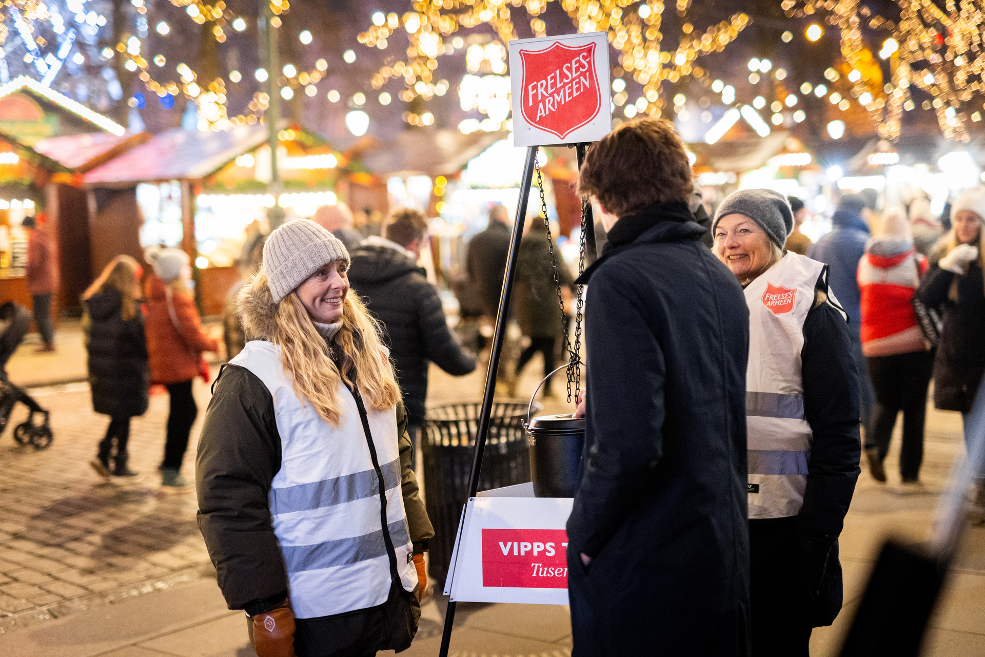 To mennesker står vakt ved julegryta. De ser på hverandre og er glade. I bakgrunnen er det masse julelys og ved siden av dem spiller en trompetist.