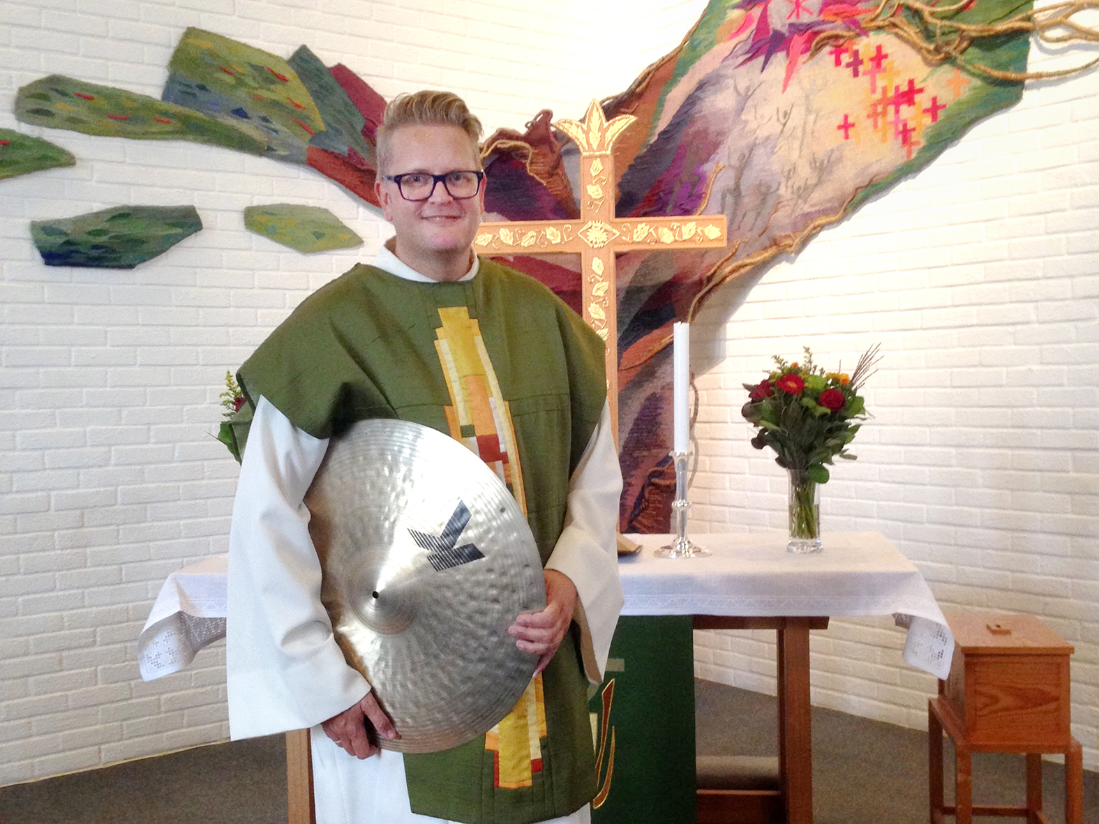 Foto: Mann i prestekjole i hvitt og grønt står foran et kors i en kirke. Under armen holder han en Hi-hat til et trommesett.