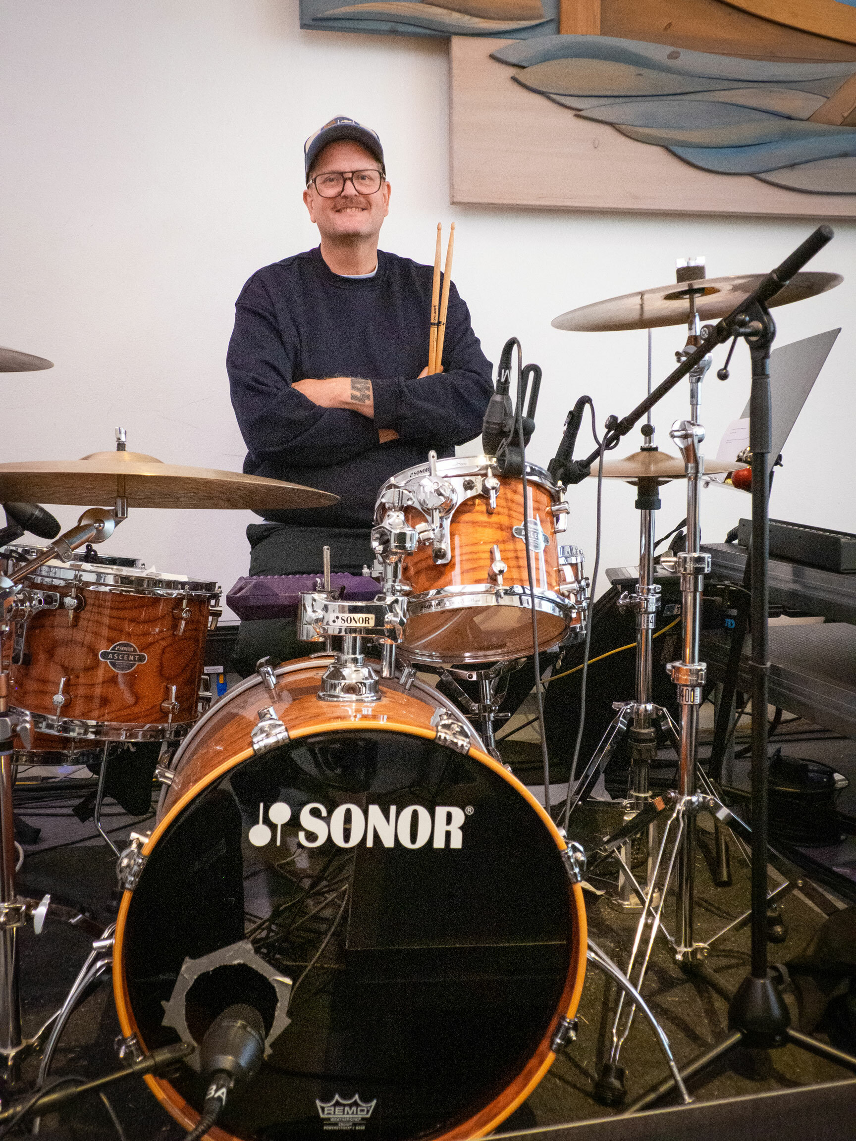 Foto: Mann i 40-årene med firkantede, store briller og caps sitter bak et slagverk. Han smiler til kamera og har trommestikkene i armene som ligger i kross.