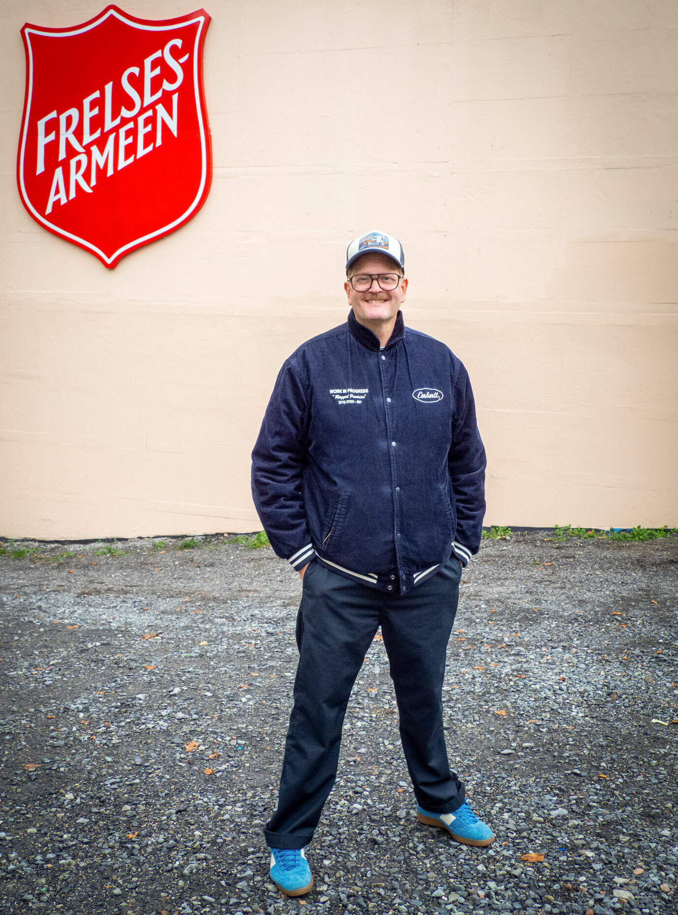 Foto: Mann i blå college jakke med hvite striper på bordene står med caps og briller utenfor et digert skilt hos Frelsesarmeen.