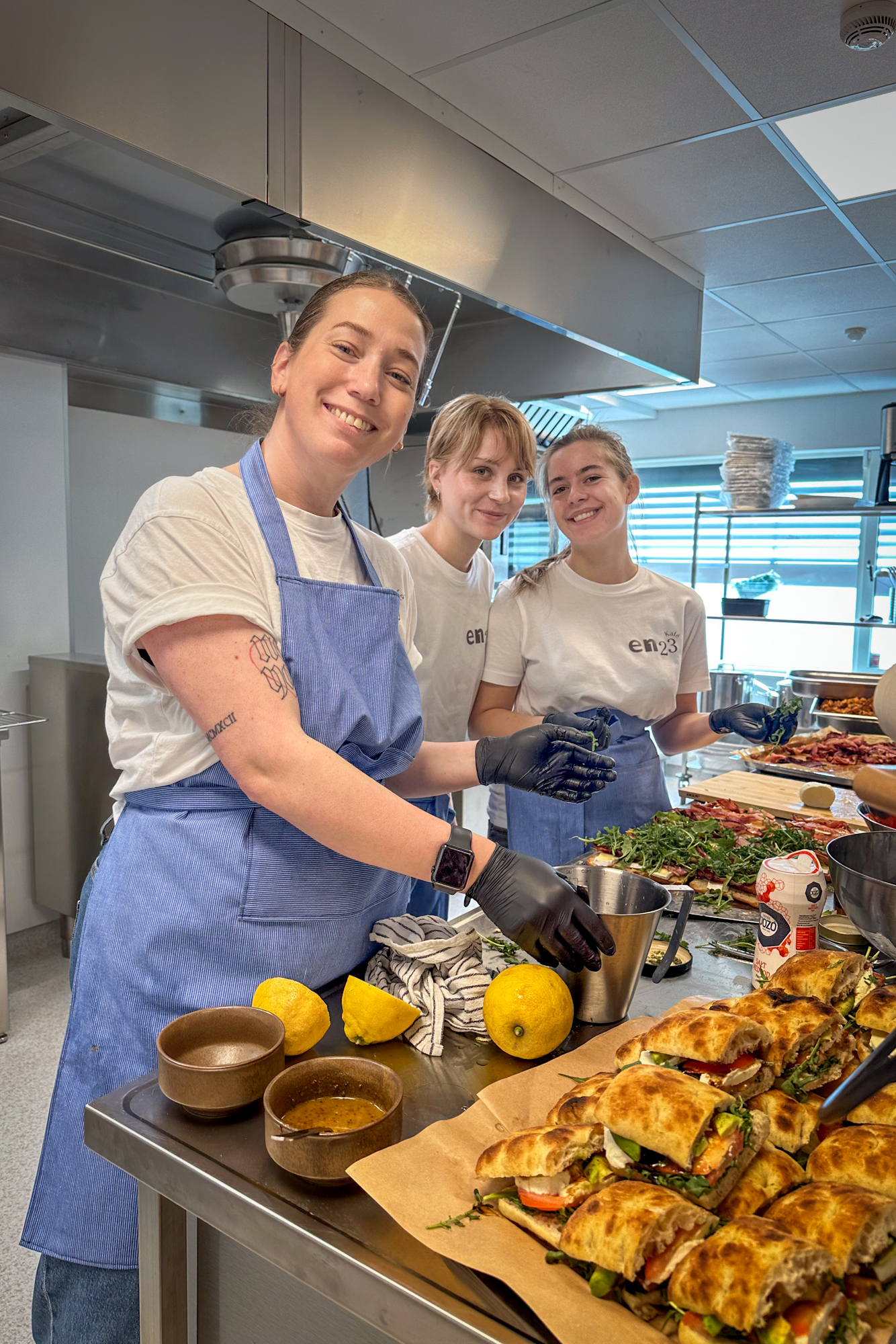 Foto: Tre damer mellom 20 og 35 år smører mat på kjøkkenet. de har på seg hvite T-skjorter med logoen til Kafe En23 og blå forklær.