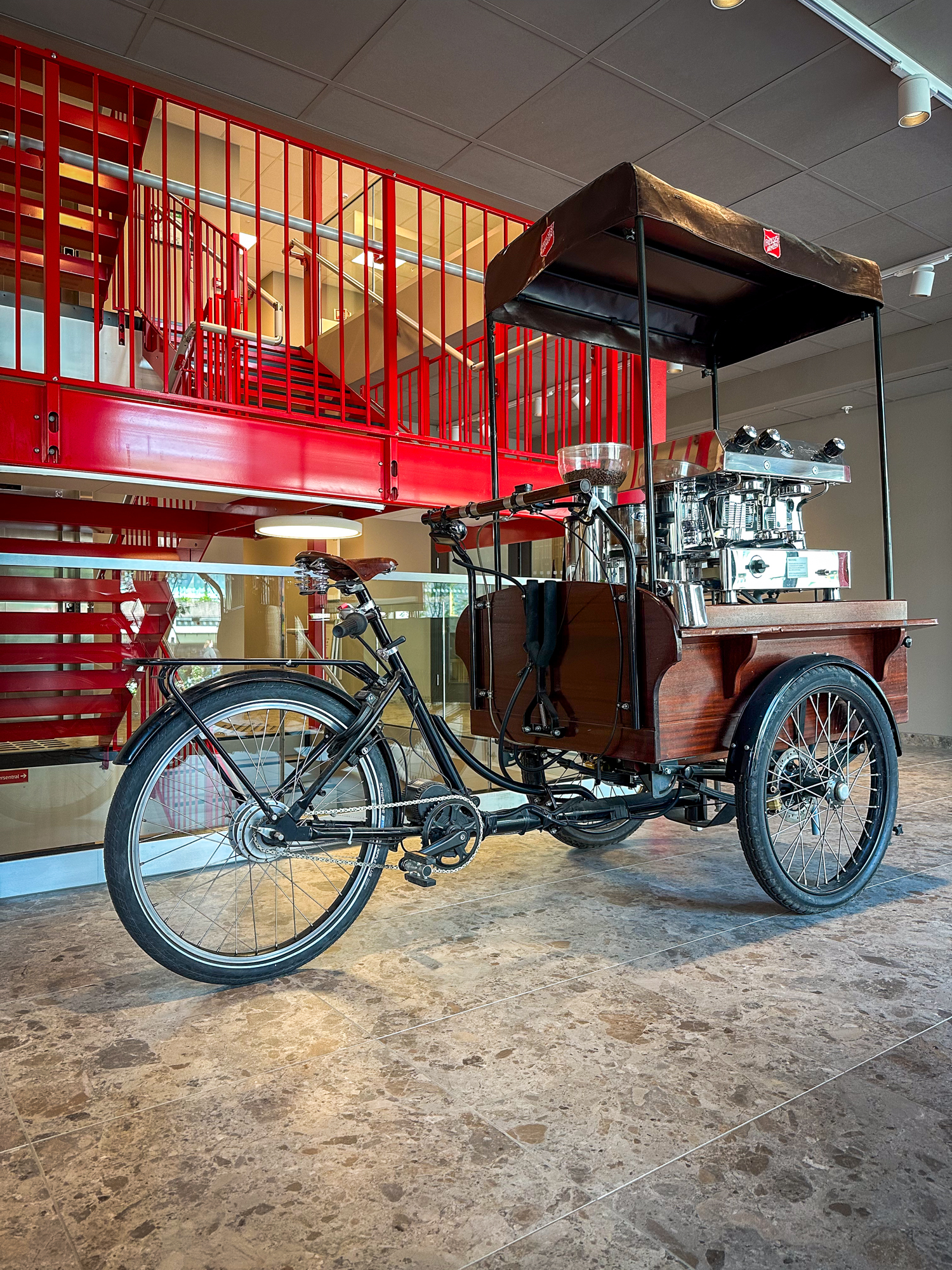 Foto: En bsort og brun sykkel med tre hjul står parkert inne på gulvet, foran en rød trapp i metall. Sykkelen har påmontert en fancy kaffemaskin foran. Over det hele er et tak i brunt skinn med logoen til Frelsesarmeen på hver side.