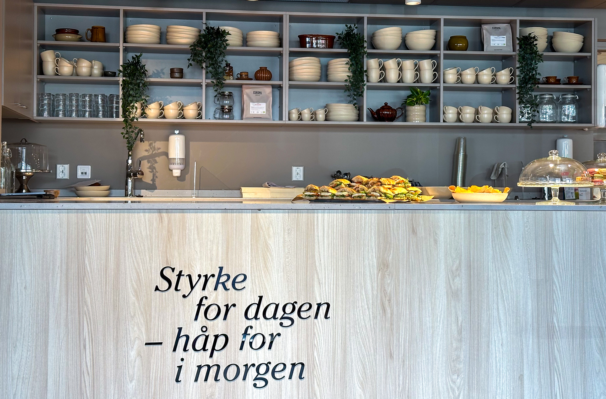 Foto: Av kafédisken med alle hyller bak, fulle av kopper, glass og skåler. Foran på det lyse treverket i disken står det en tekst.