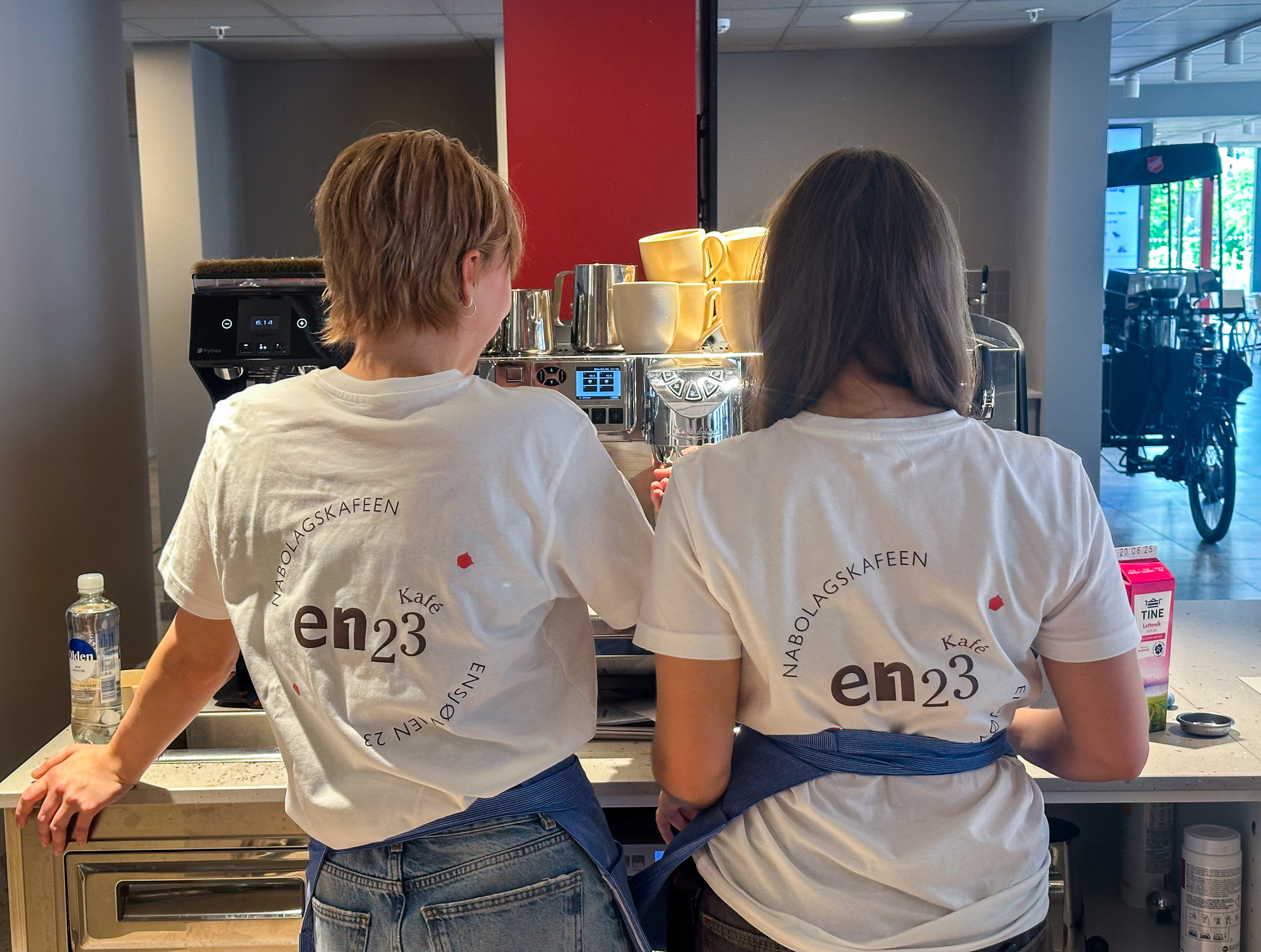 Foto: To unge kvinner står med ryggen til kaffemaskinen. De har hvite T-skjorter på der det står Kafé En23.