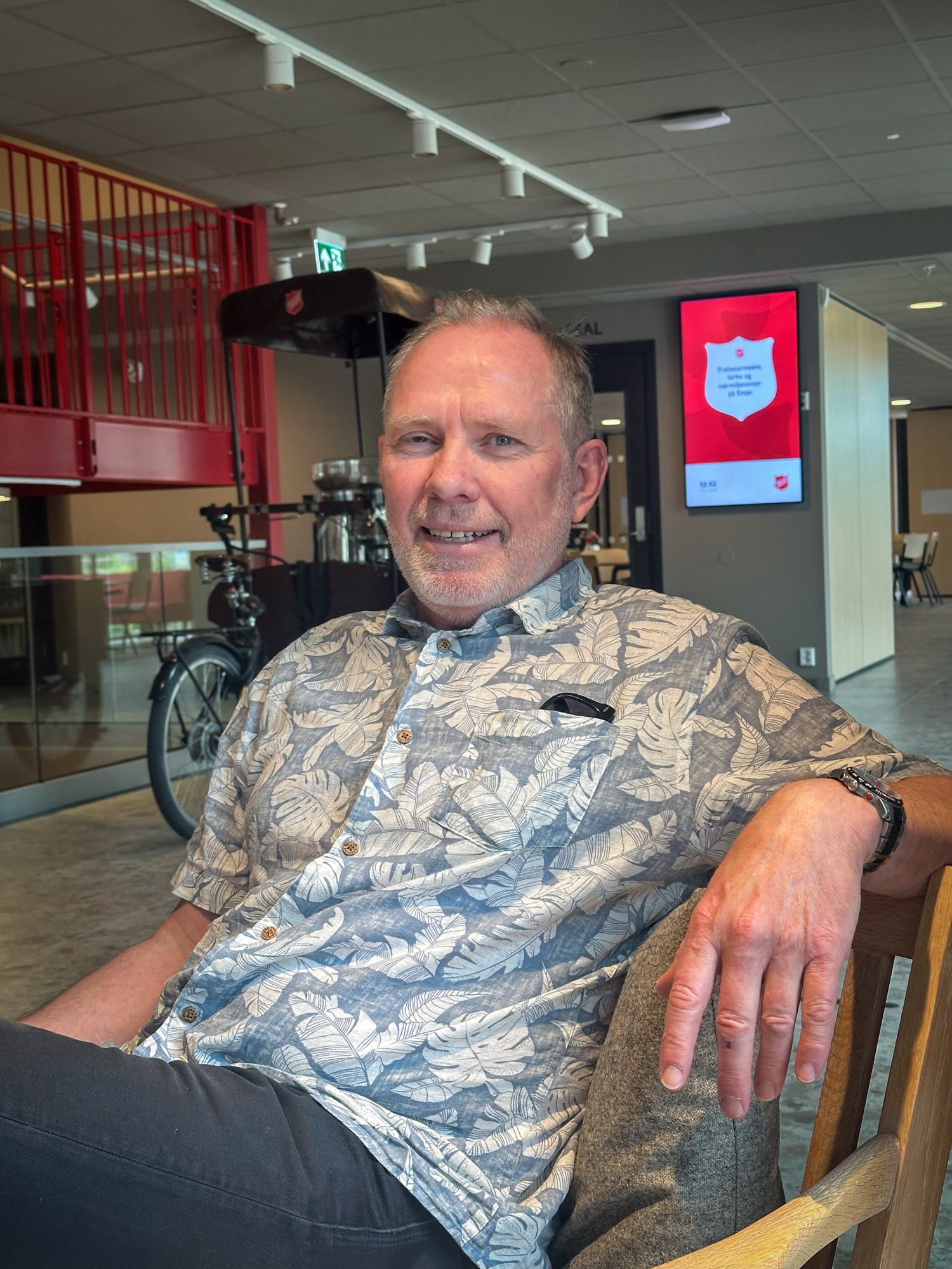 Foto: Mann i blomstrete skjorte i blått og hvitt sitter henslengt i en stol. Bak ham på en informasjonsskjerm sees Frelsesarmeens skjold. Det står også en sykkel der med kaffemaskin installert foran på sykkelen. Doningen har tak over seg med logoen til Frelsesarmeen..